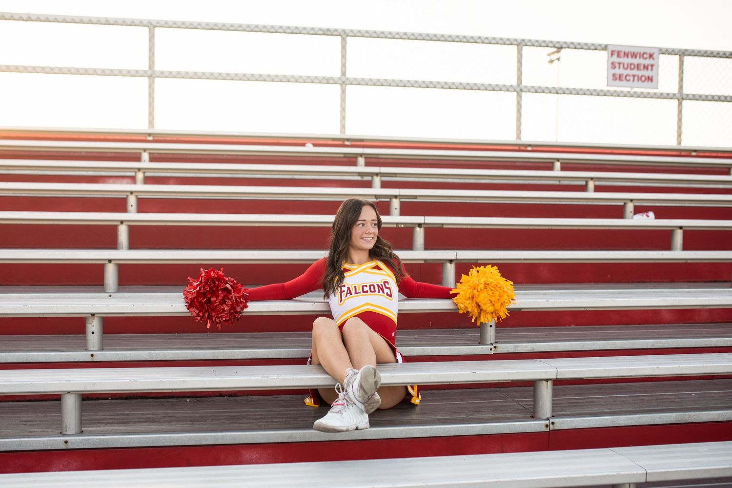 A cheerleader in white uniform sits on red bleachers holding orange and red pom poms at a sports stadium.