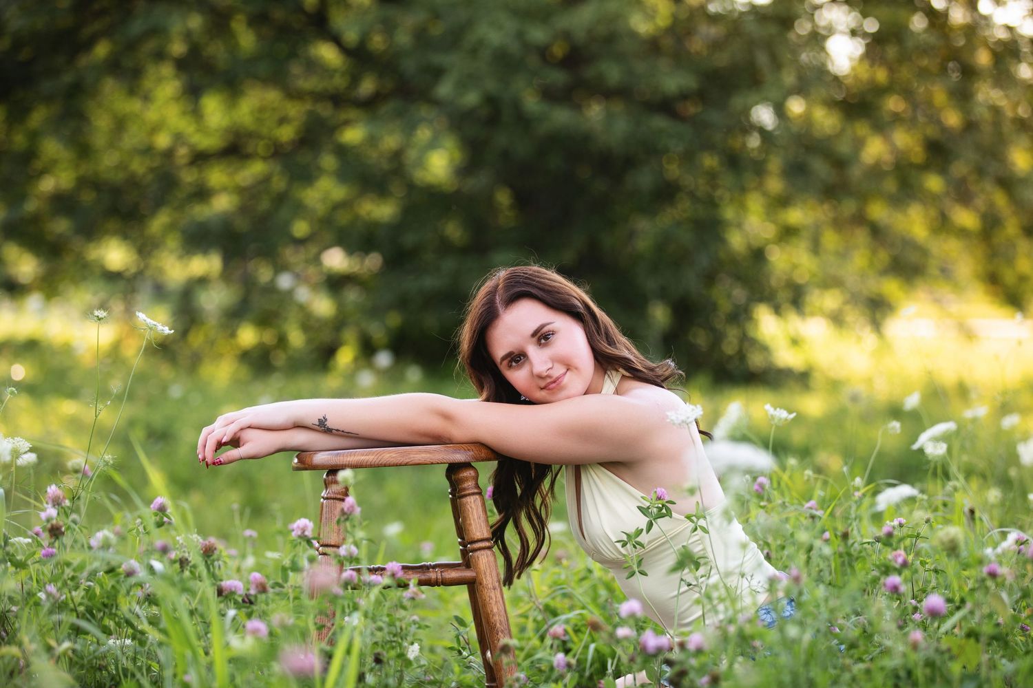 Young woman in white dress leaning on wooden chair surrounded by wildflowers in a lush green meadow.