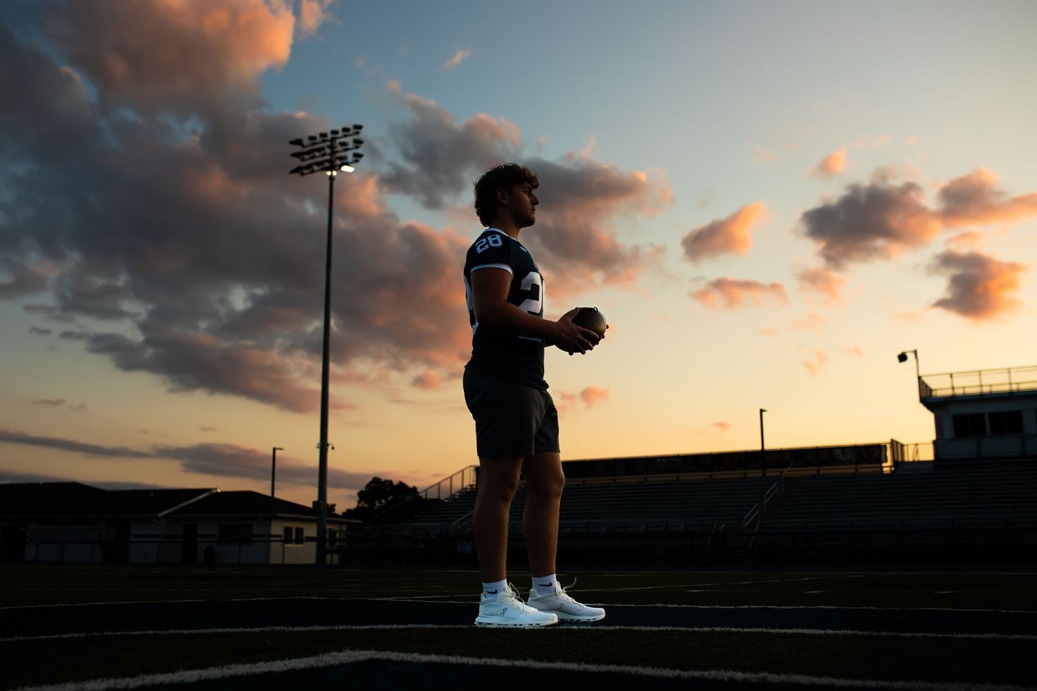 A silhouetted basketball player stands against a dramatic sunset sky at an outdoor court.