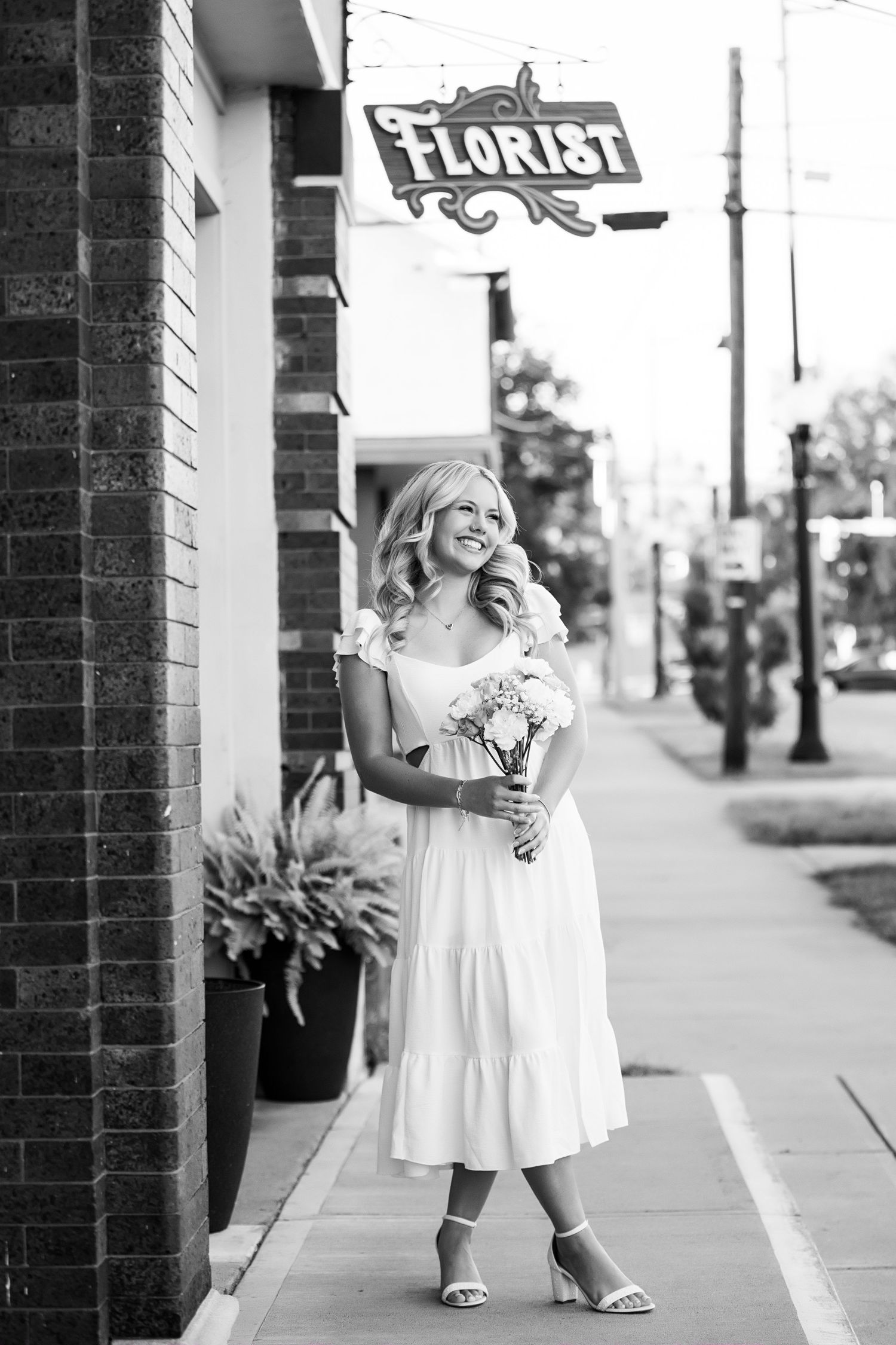 A black and white photo series shows a person in a white dress posing on a brick-lined sidewalk near a florist shop.