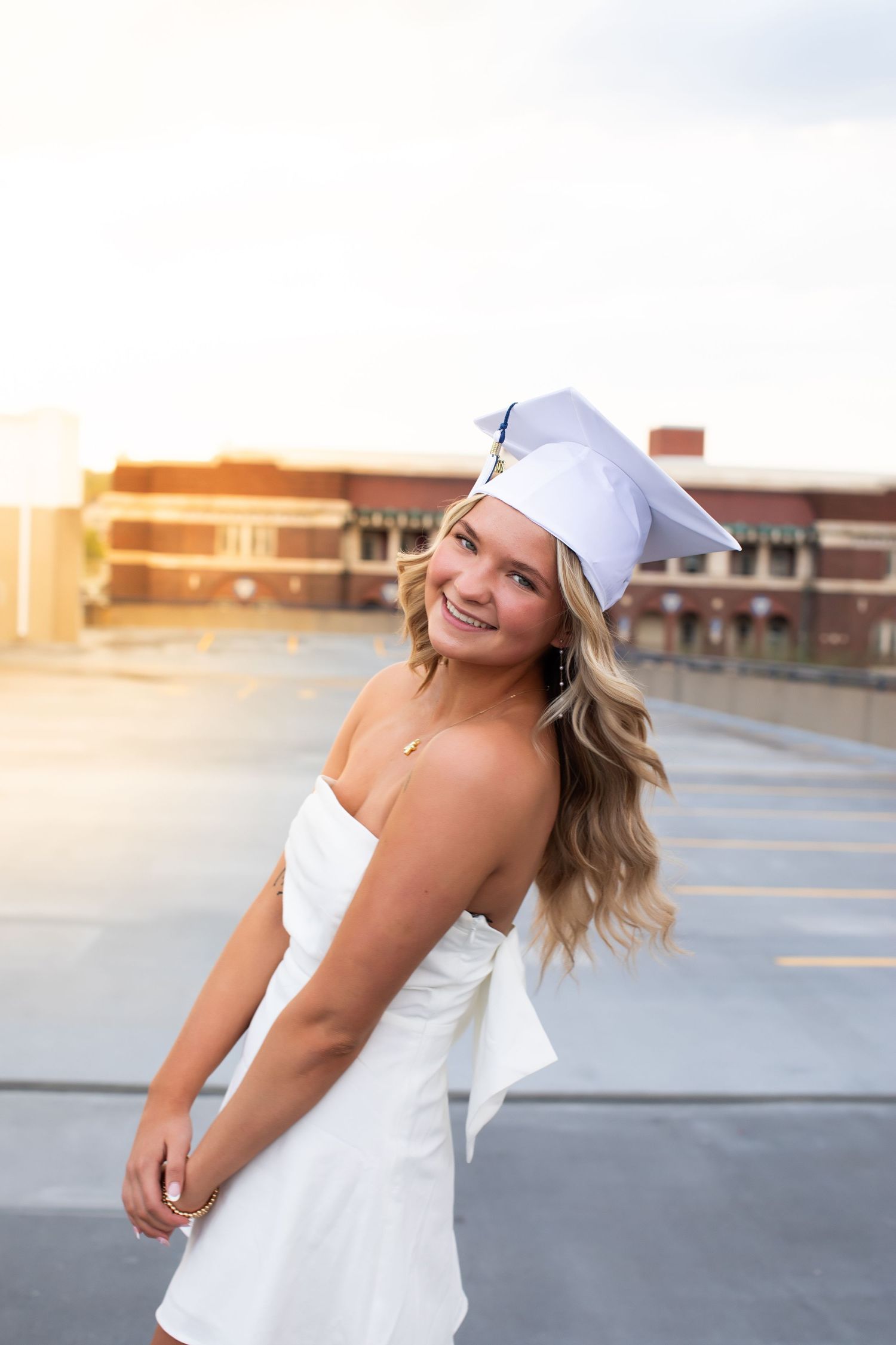 A person in a white dress and baseball cap poses playfully in a parking lot at sunset.
