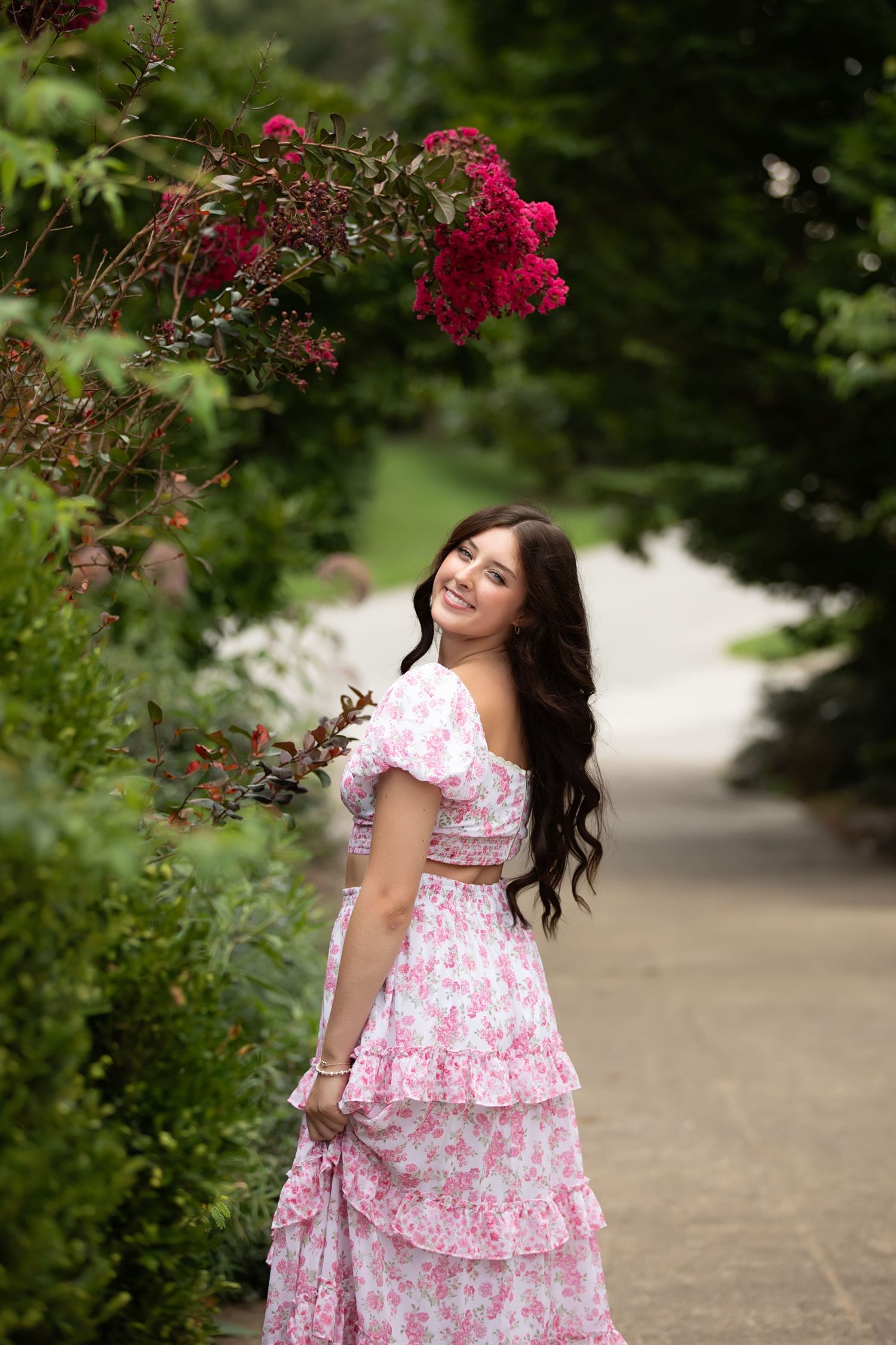 A figure in a pink floral dress walks along a garden path surrounded by vibrant red roses and greenery.