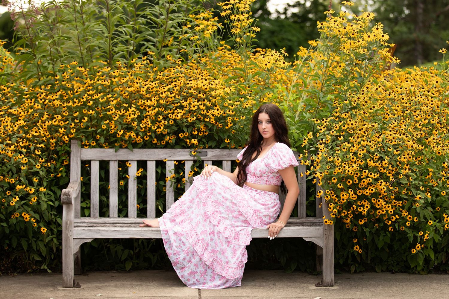 A person in a pink floral dress poses gracefully on a wooden bench surrounded by vibrant yellow wildflowers.