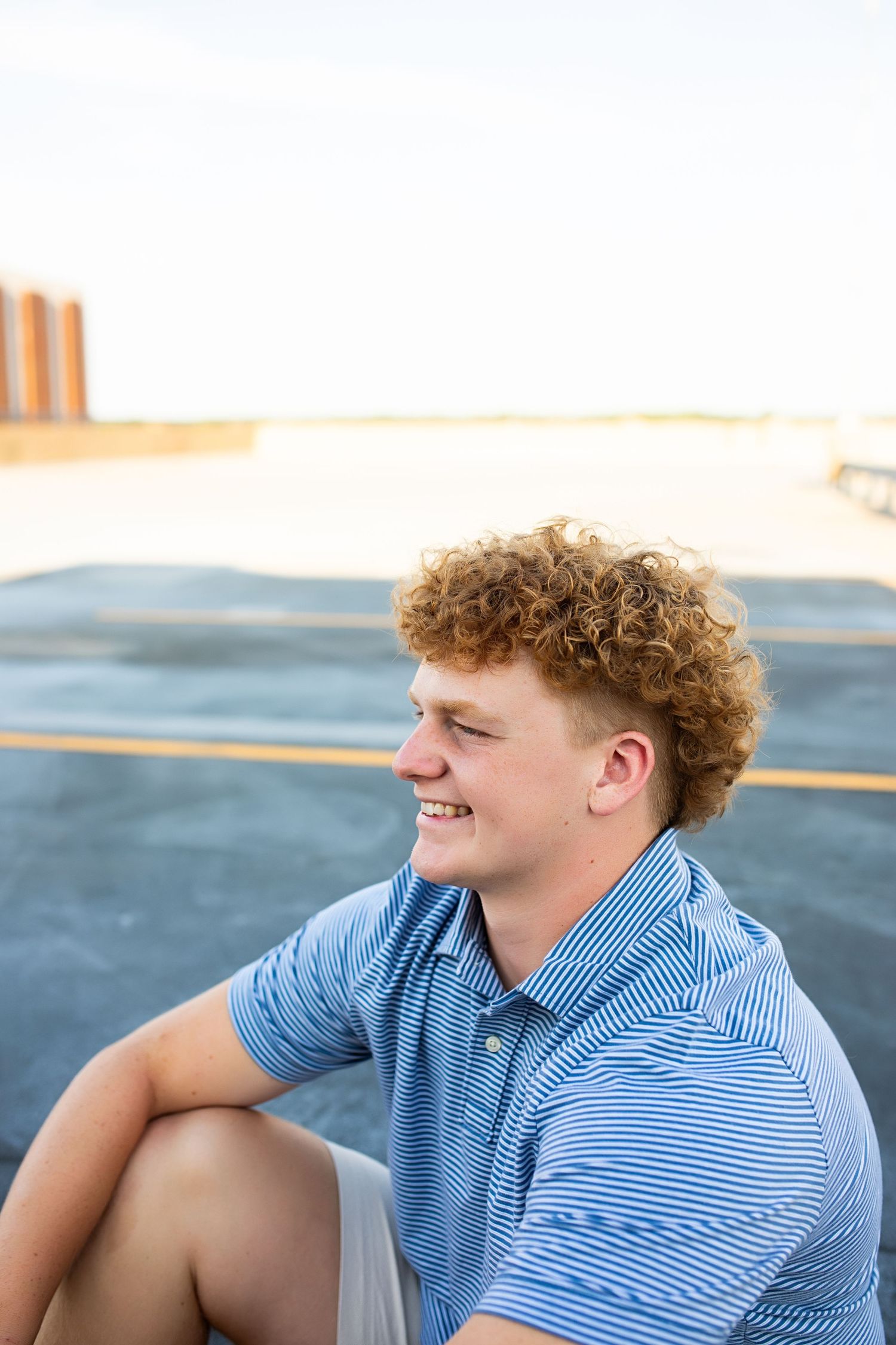 A laughing person in a blue striped polo shirt sits on a sandy beach near the ocean on a sunny day.