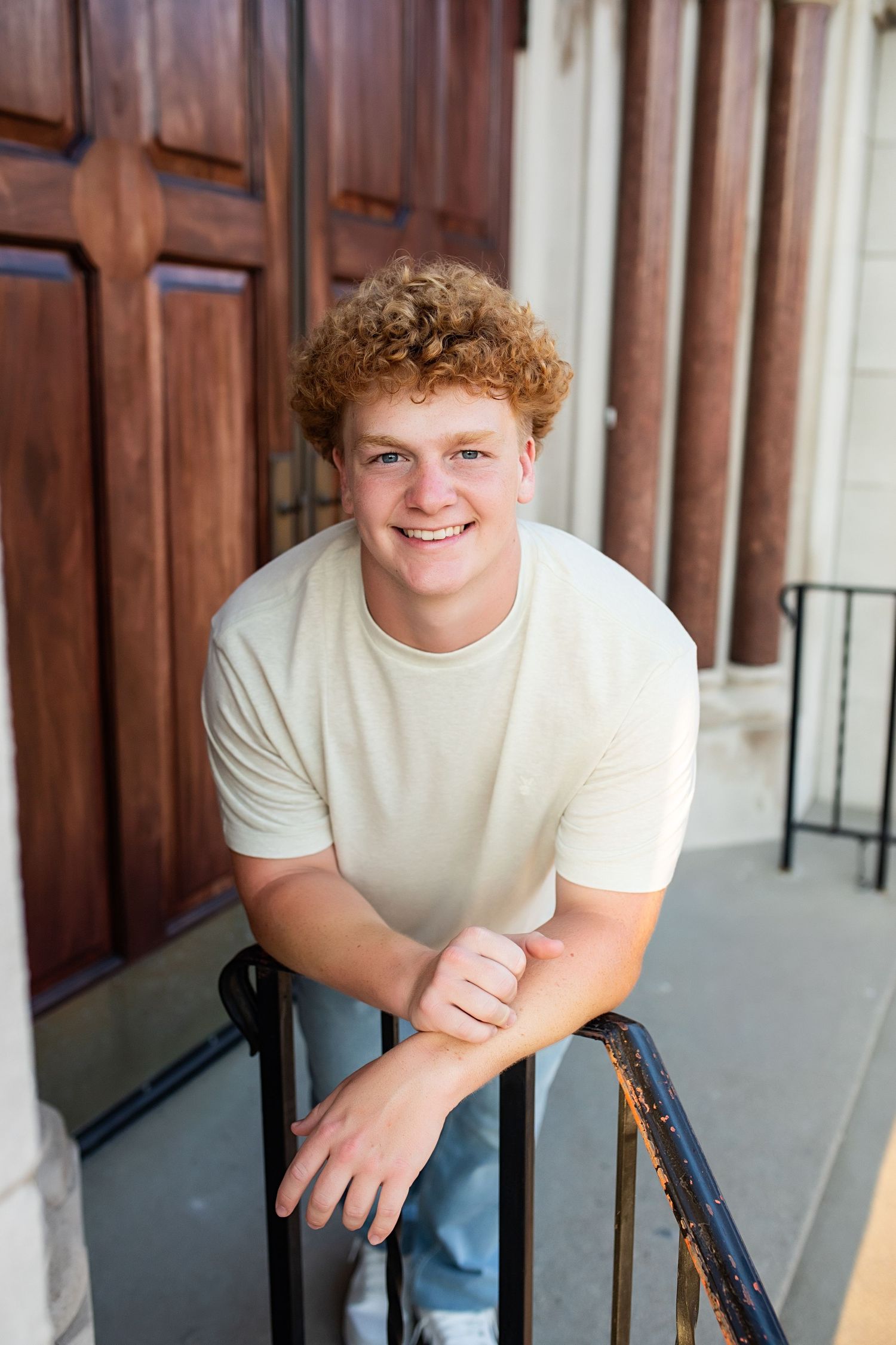 Young person in white t-shirt sits by wooden panels smiling at camera in outdoor portrait setting.