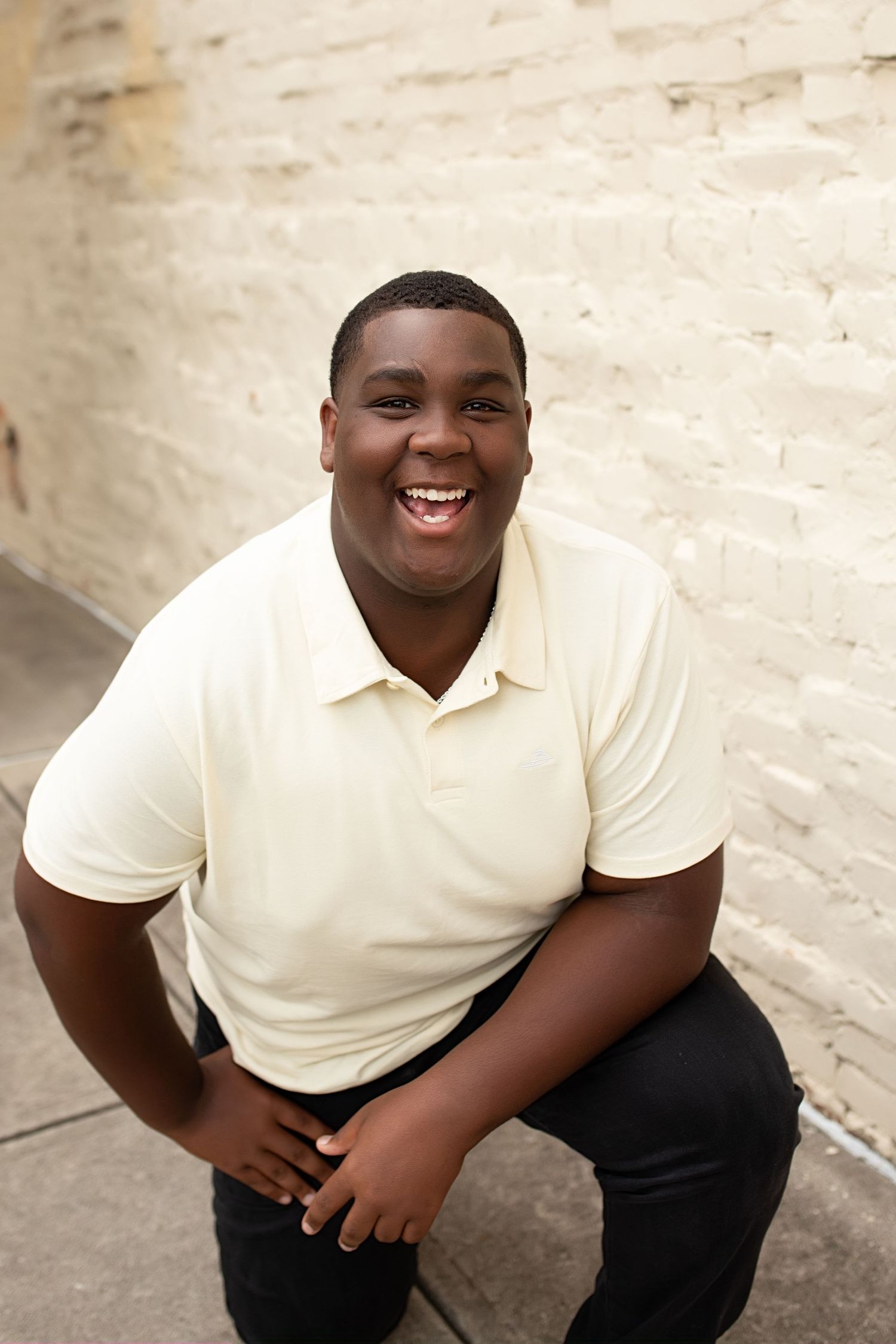 A person wearing a cream colored polo shirt and dark pants smiles warmly while sitting against a light-colored wall.