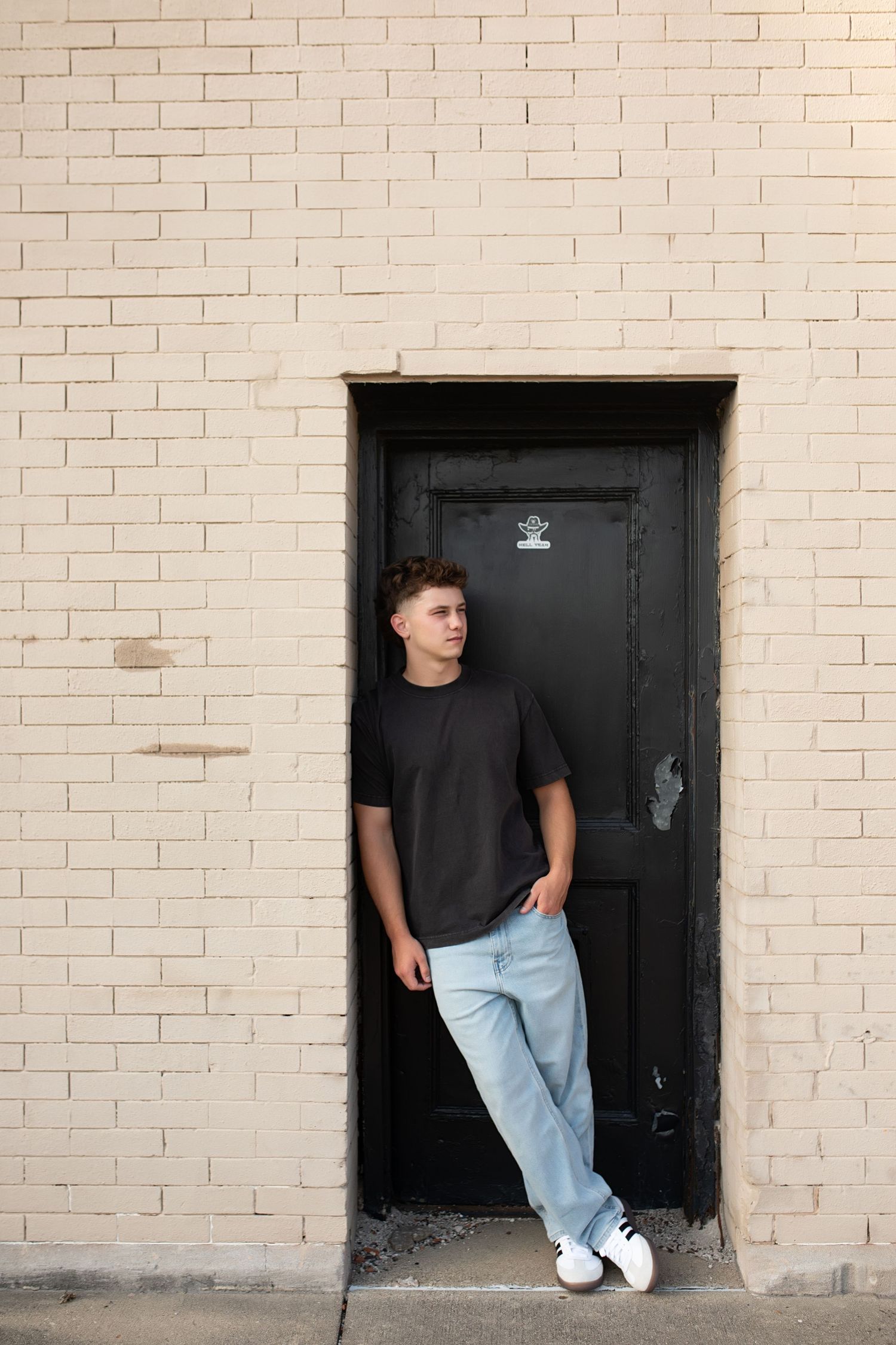 Person in a black t-shirt and light jeans leaning against a doorway in a brick wall.