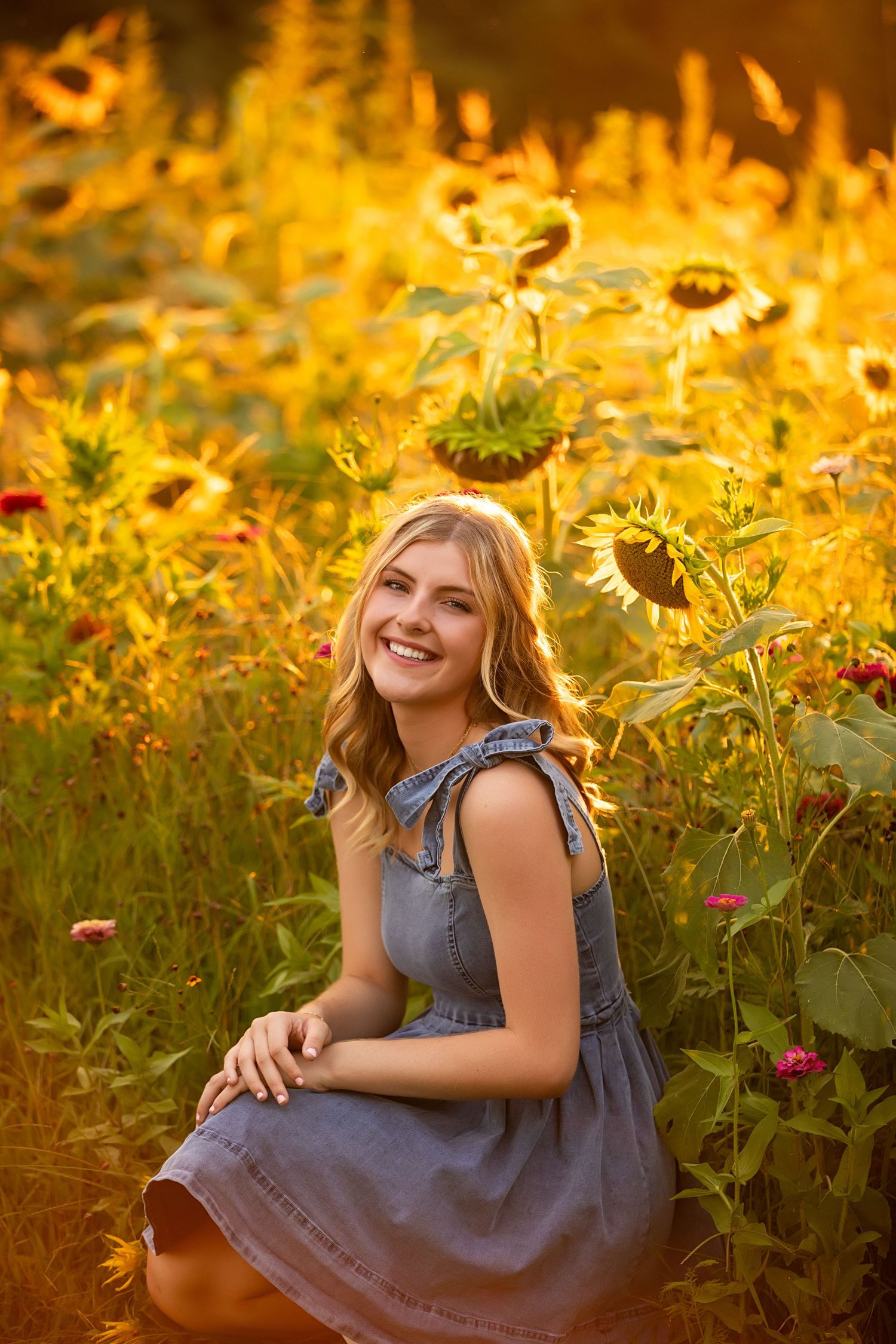 A person in a denim dress poses joyfully among golden sunflowers during sunset in a flower field.