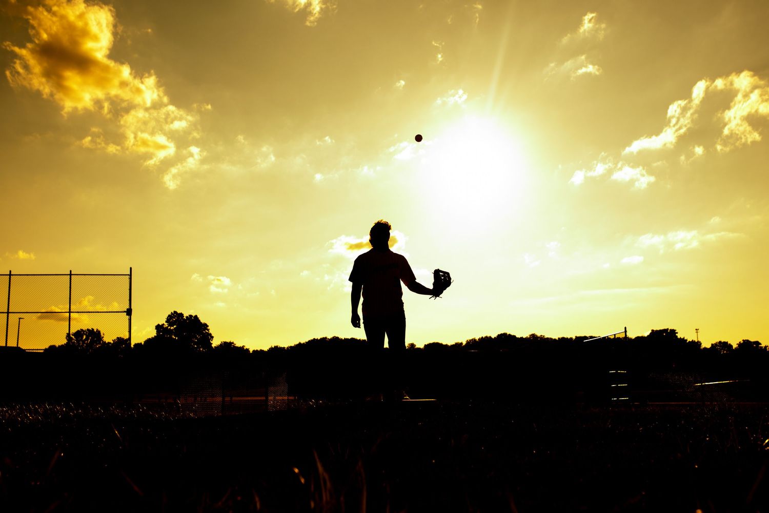 A silhouetted figure stands beside a fence in front of a dramatic golden sunset casting a warm glow over the landscape.