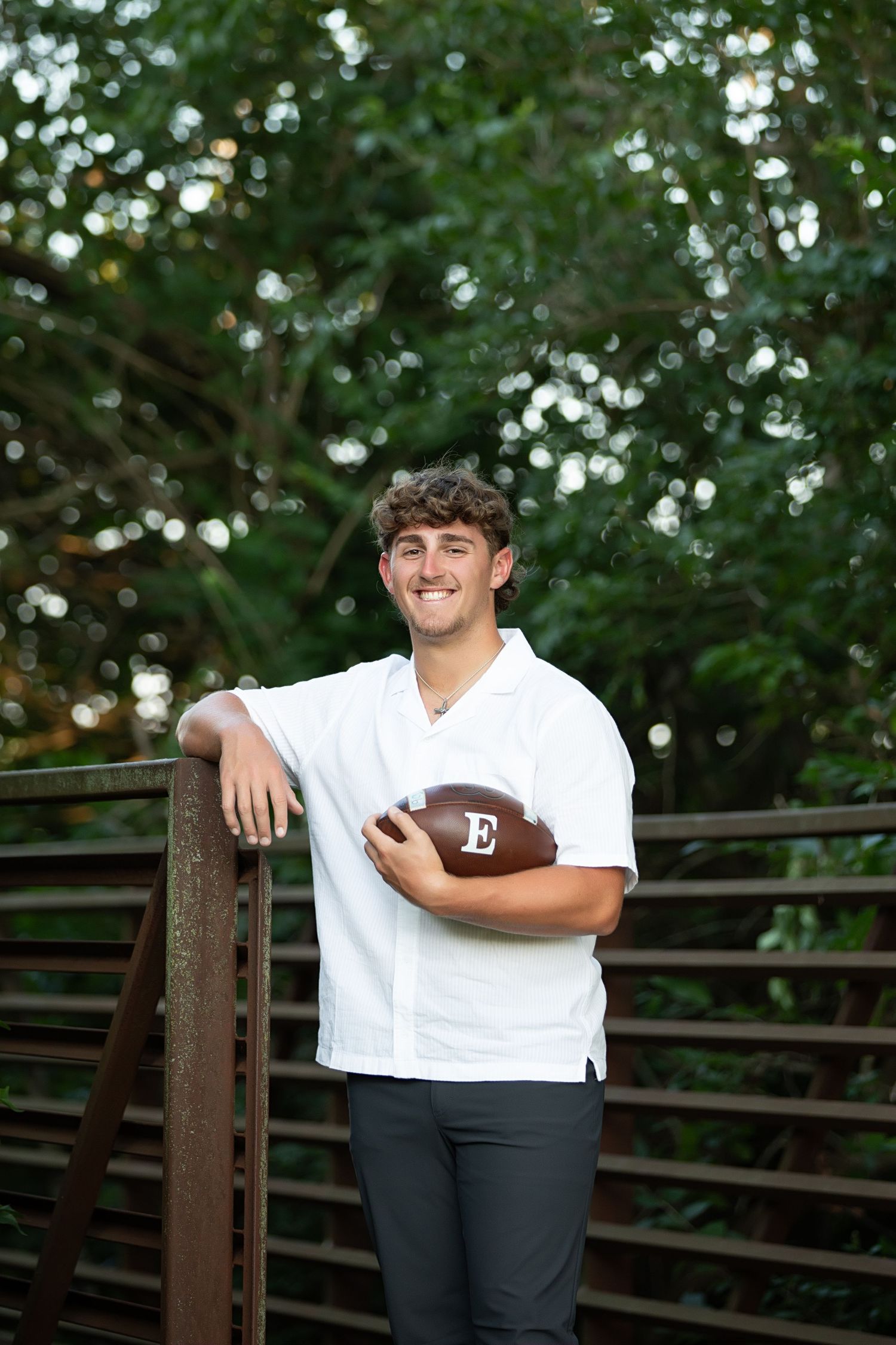 A high school football player wearing a white jersey and holding a football poses against a wooden fence.