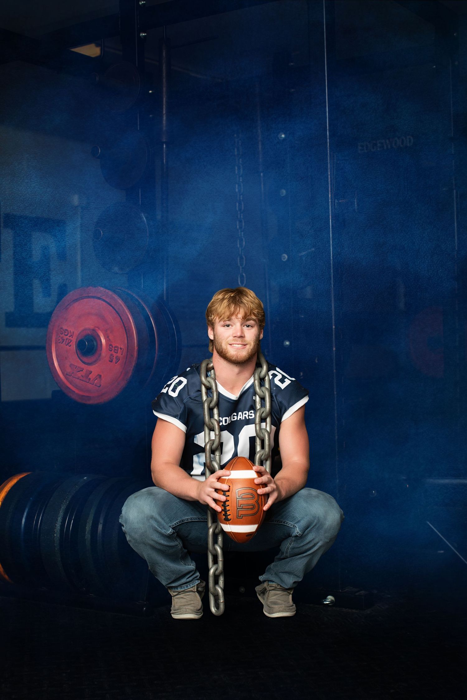A football player in a dark jersey sits on a bench holding a football against a dramatic blue-lit background.