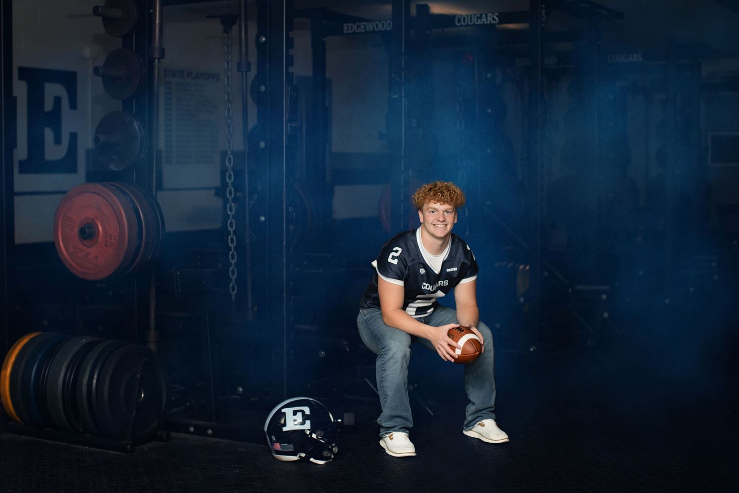 Athlete rests on weight lifting equipment in dimly lit gym with moody blue lighting and stacked plates in background.