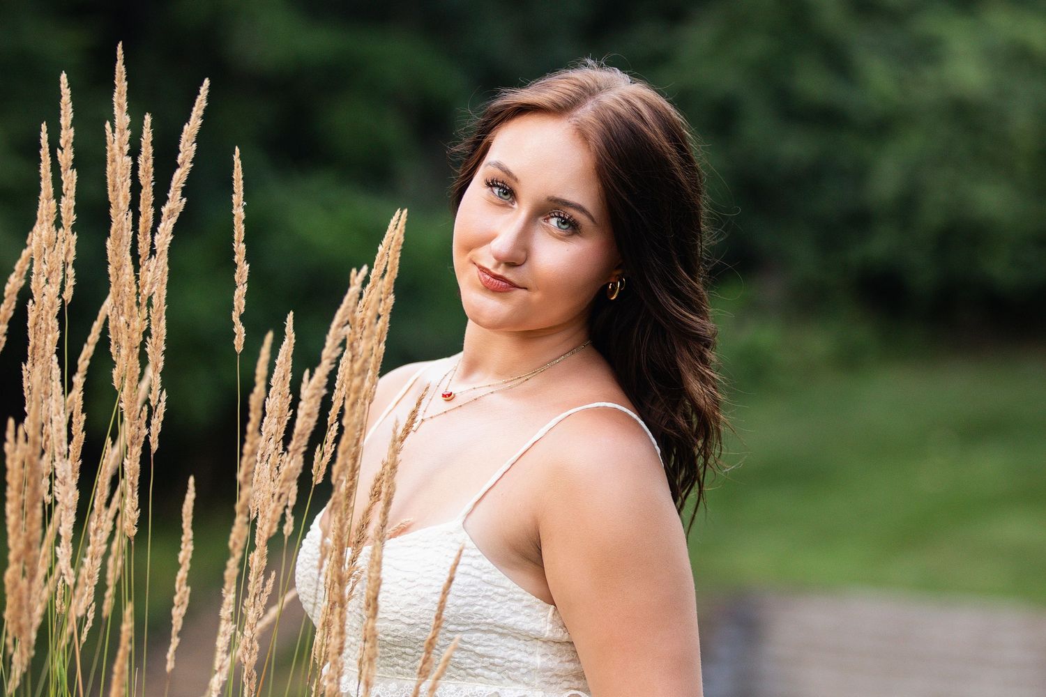 Young woman in white dress poses near golden wheat grass in a lush green outdoor setting.