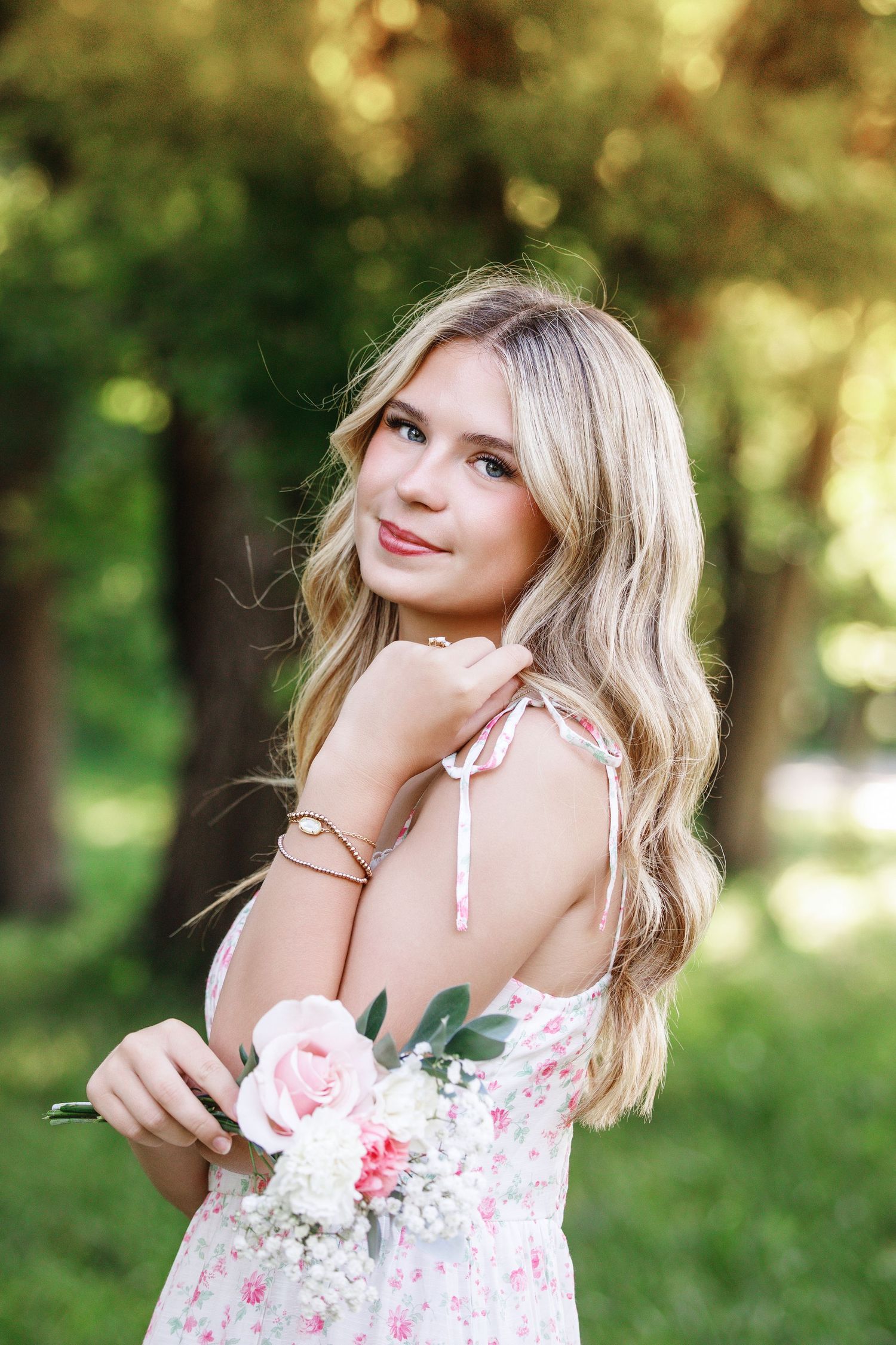 Young blonde woman in floral dress holding pink roses, smiling outdoors in a lush green park setting.