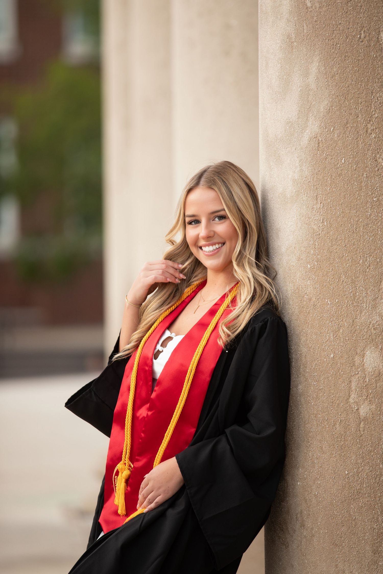 Smiling blonde woman in black graduation gown with red and gold sash leaning against a stone column outdoors.