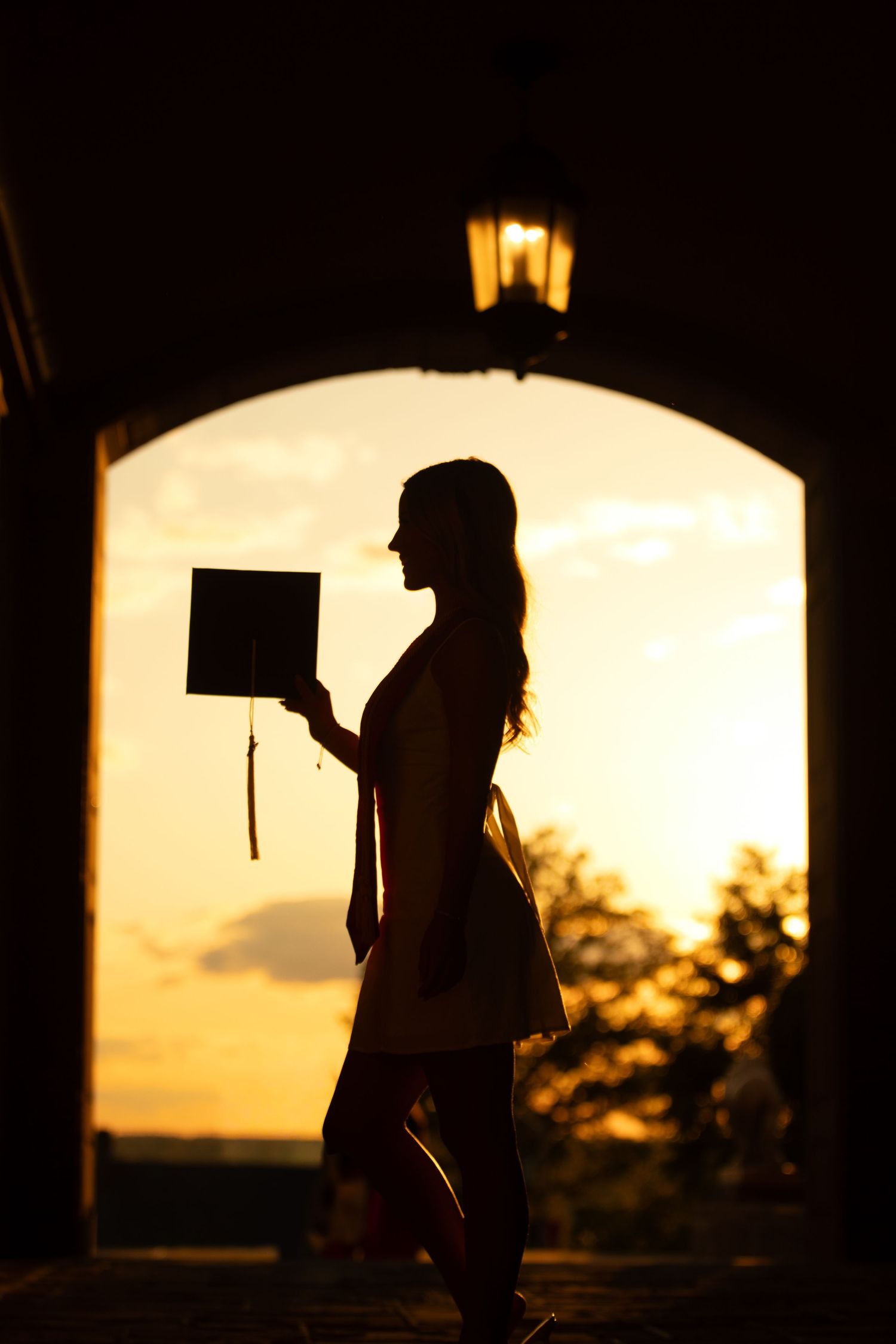Silhouette of a graduate holding a cap at sunset framed by an arched doorway with warm golden light.