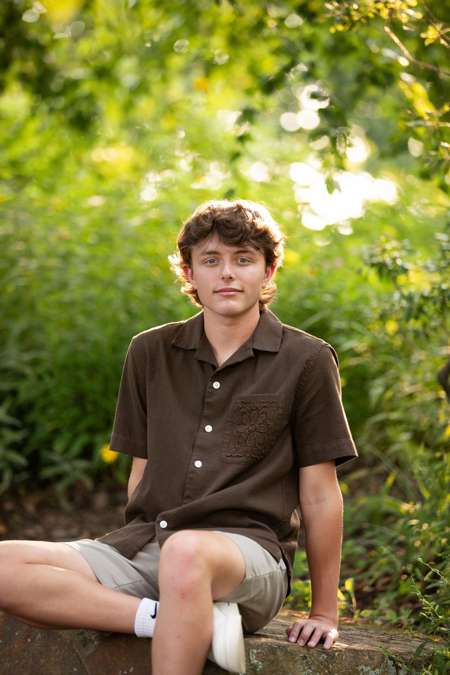Teen boy in brown shirt and khaki shorts sitting outdoors surrounded by lush green foliage.