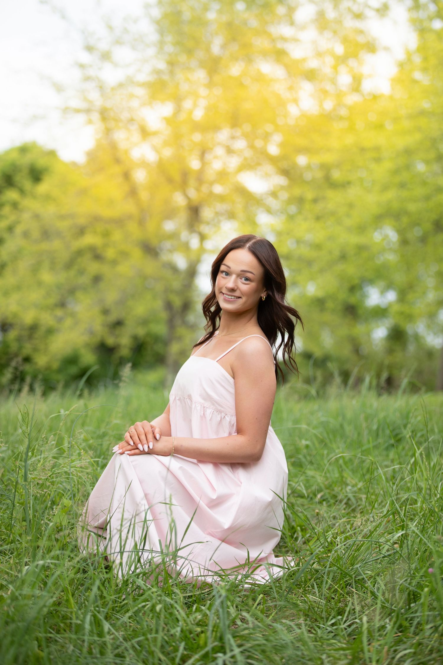 Young woman in pink sundress sitting in lush green grass, smiling outdoors with trees in background.