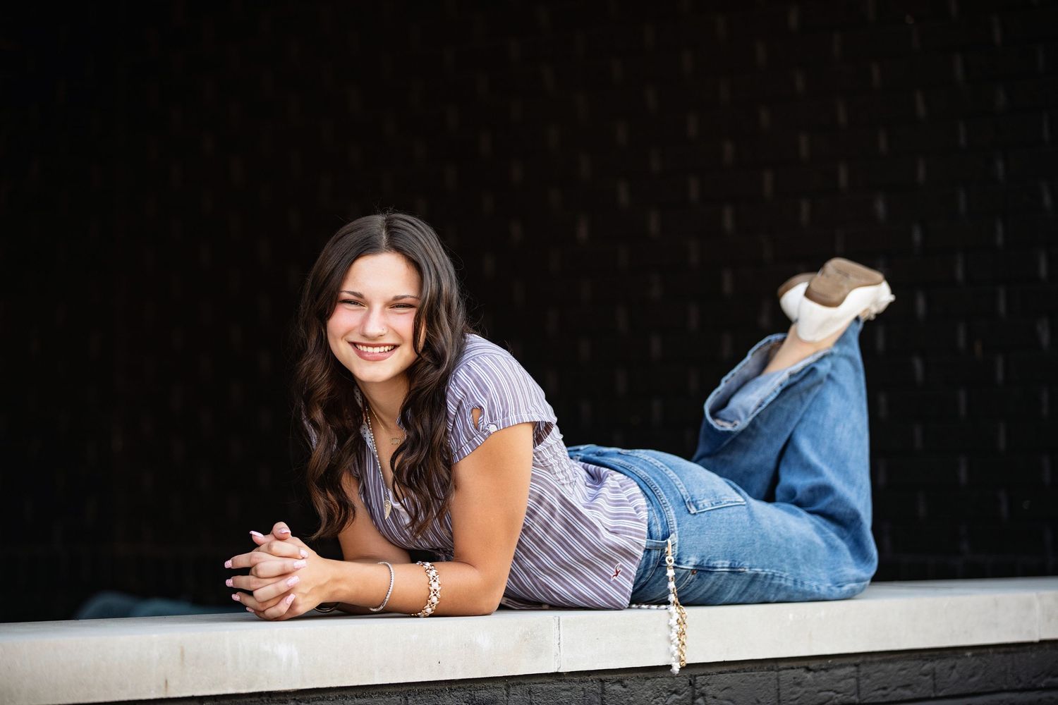 Young woman in jeans and striped top poses lying down on ledge, smiling at camera against dark background.