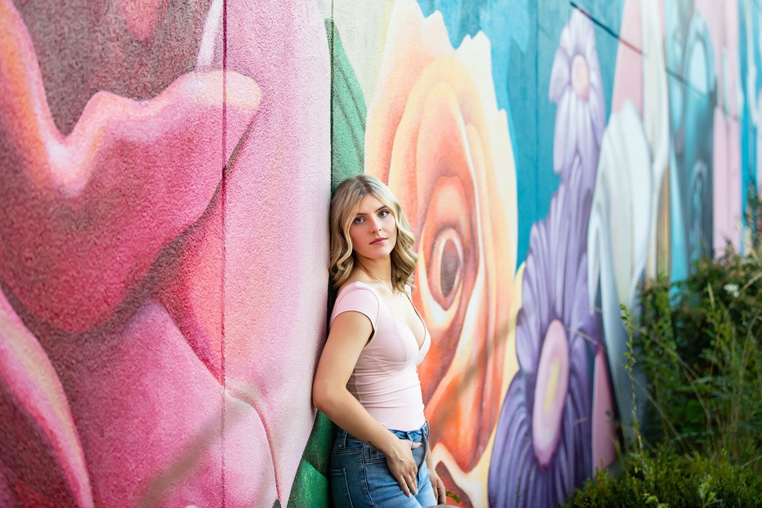 A person poses casually against a vibrant outdoor mural wall featuring pink, orange and blue abstract designs.