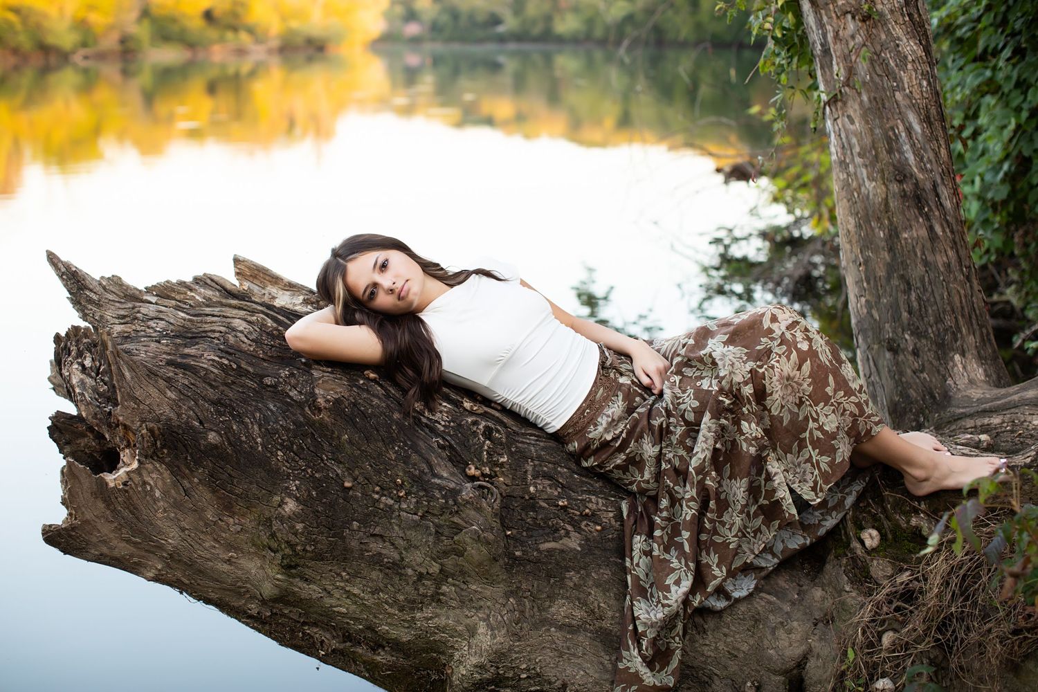 A woman in a white dress lies gracefully on a fallen tree trunk over water during a dreamy outdoor photo session.