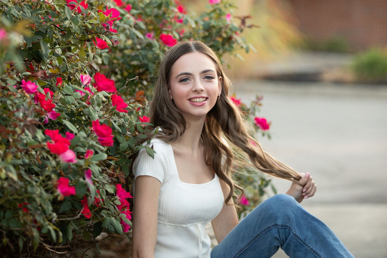 A person in a white top and jeans sits among vibrant red roses in a garden setting.