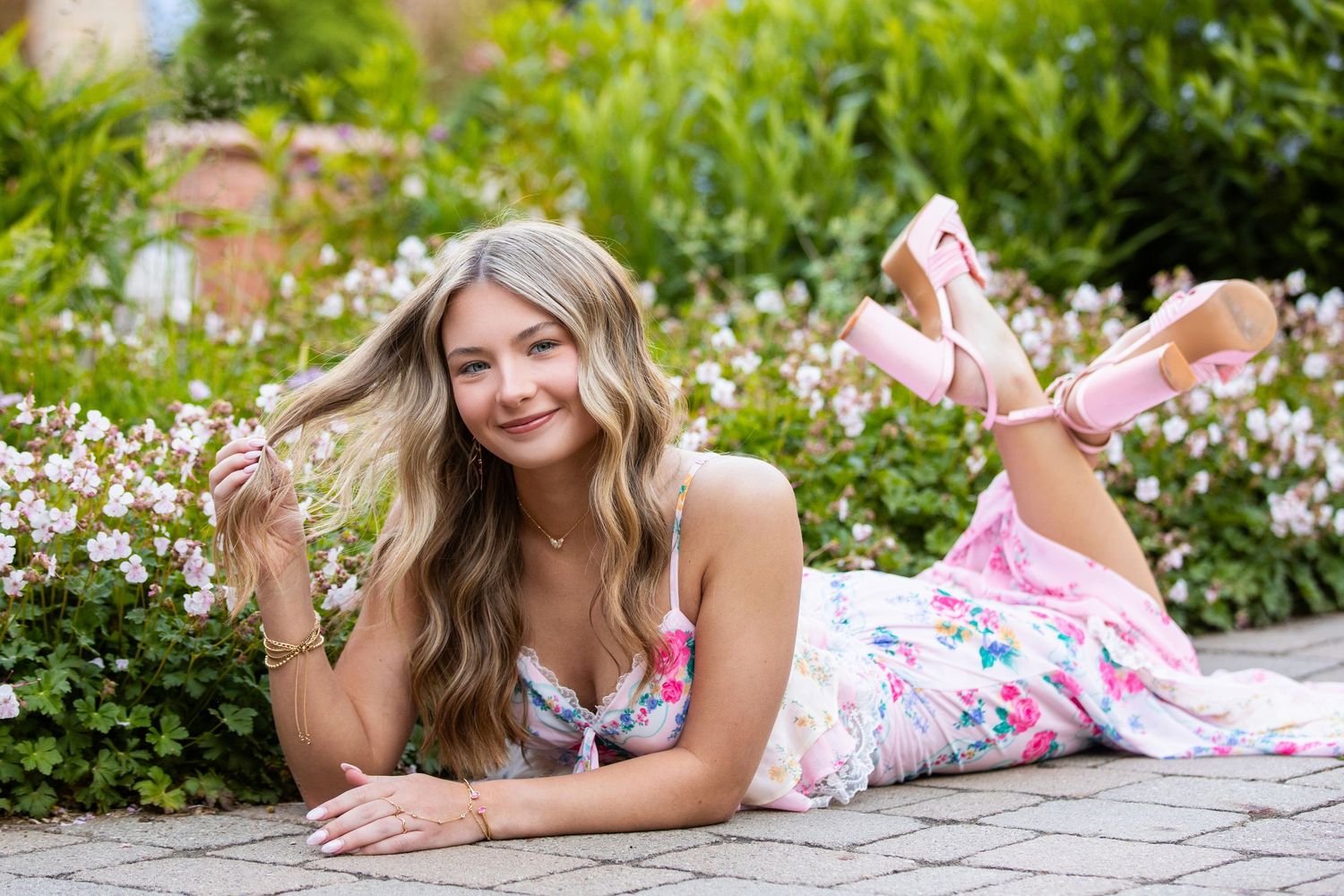 Young blonde woman in floral dress lying on garden path surrounded by blooming flowers, smiling at camera.