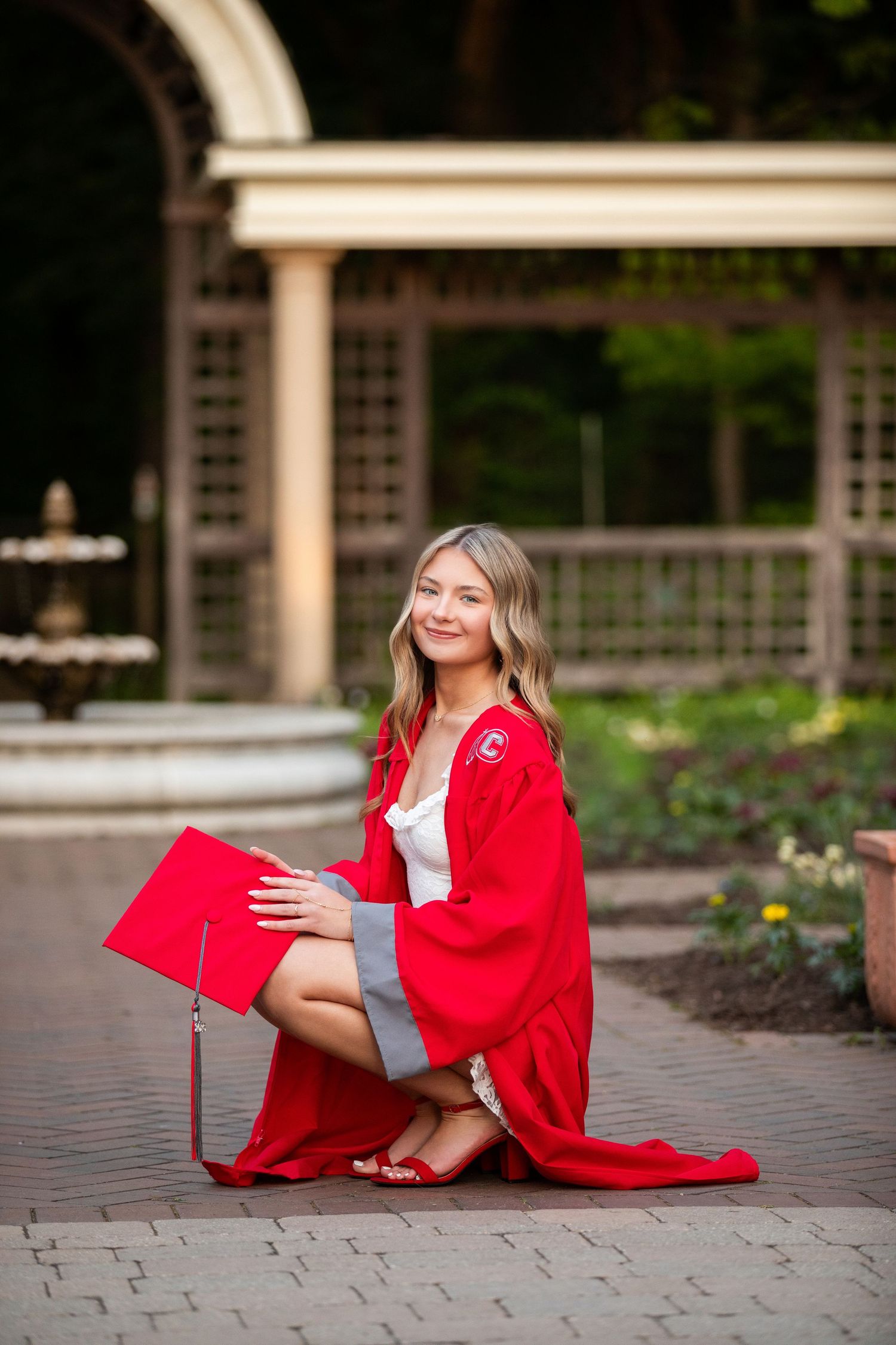 Young woman in red graduation gown and cap kneeling on brick path in garden setting, smiling at camera.
