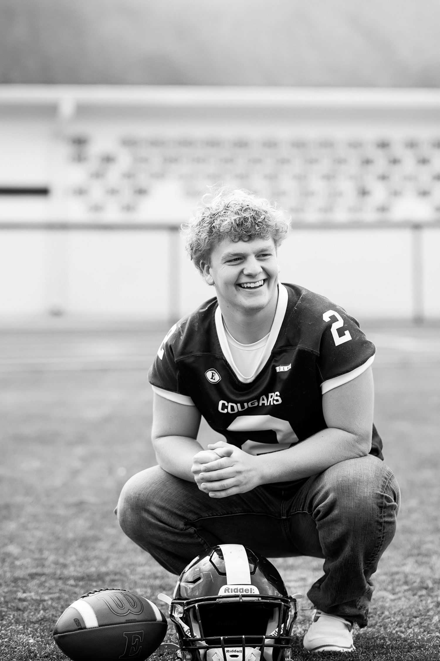 Black and white photo of a football player kneeling on field with helmet and ball in stadium setting.