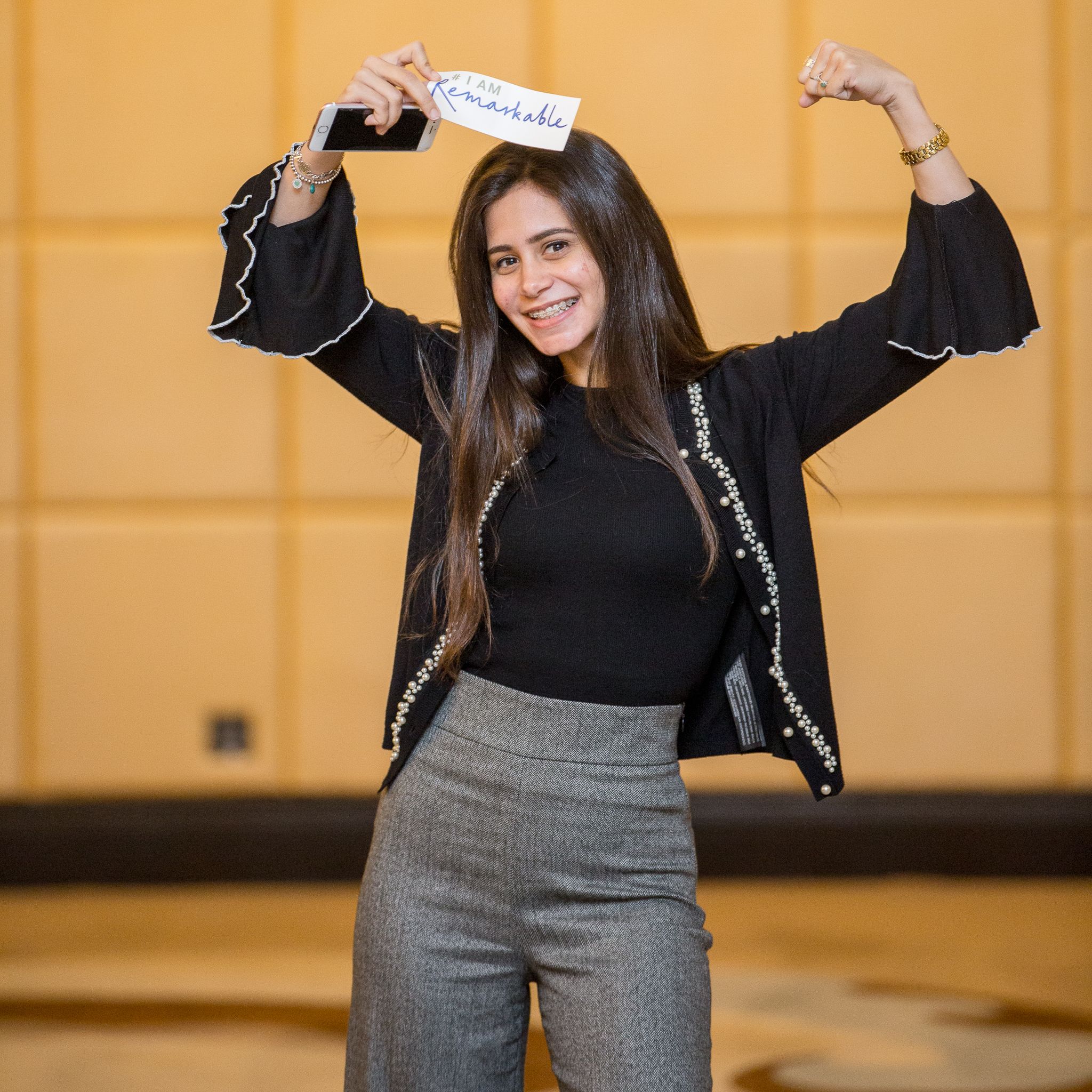 Person in black top and gray pants posing energetically against orange wall.