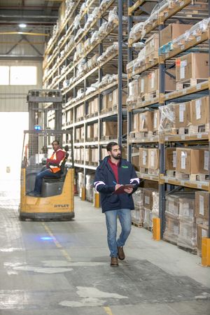 Industrial warehouse interior with forklift operator and worker checking inventory among storage racks.