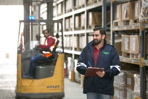 Workers checking inventory in a large warehouse with forklift and tall storage shelves.