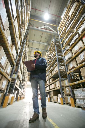 Worker with clipboard examining inventory in tall warehouse shelving aisle.