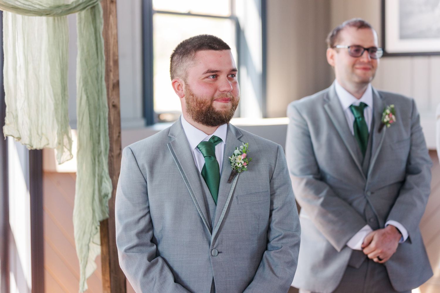 Two groomsmen in gray suits with emerald green ties stand at the altar in a bright indoor ceremony space.