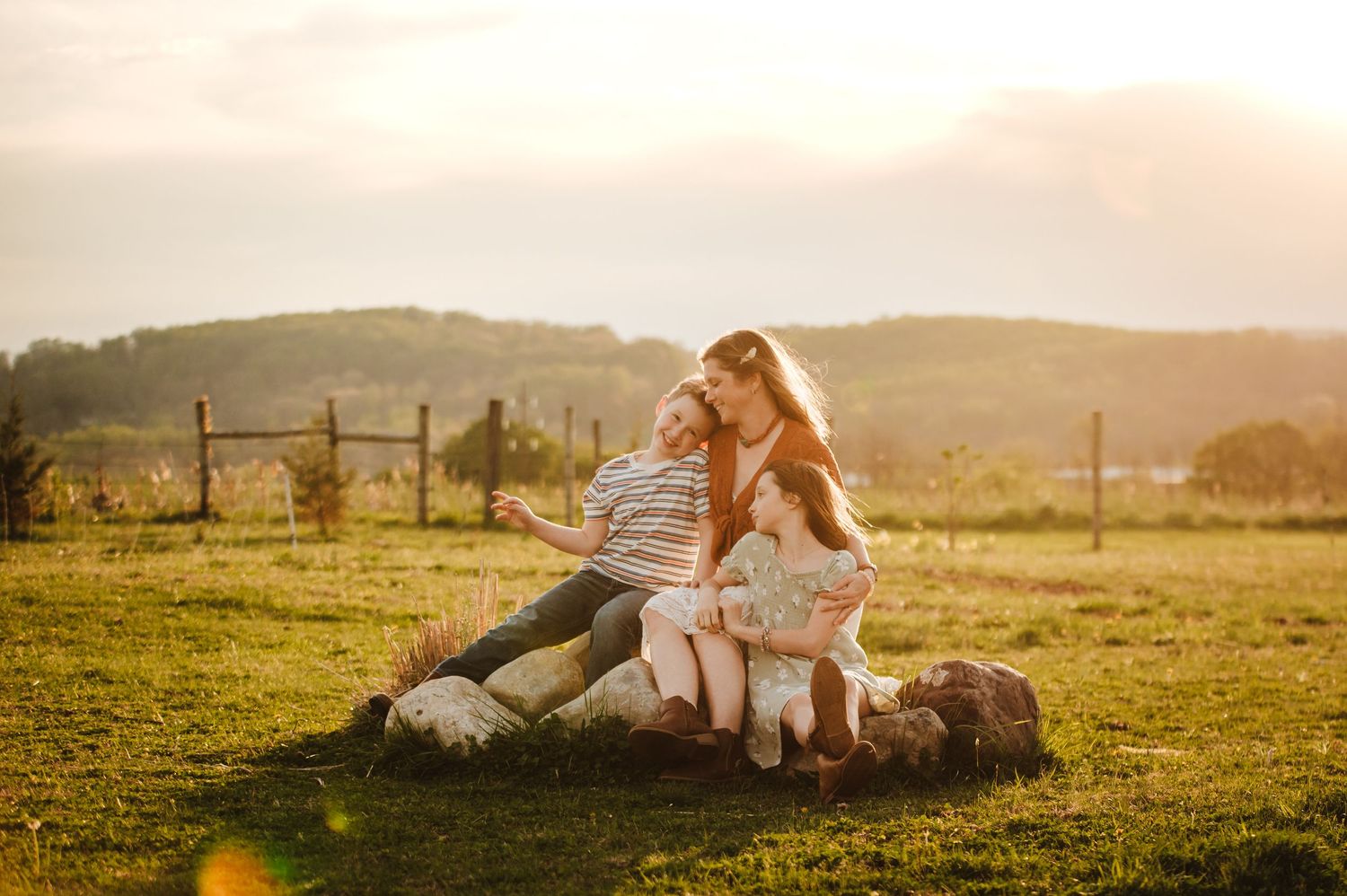 A mother sits on rocks with her two children in a sunny green field at golden hour, sharing a tender family moment.