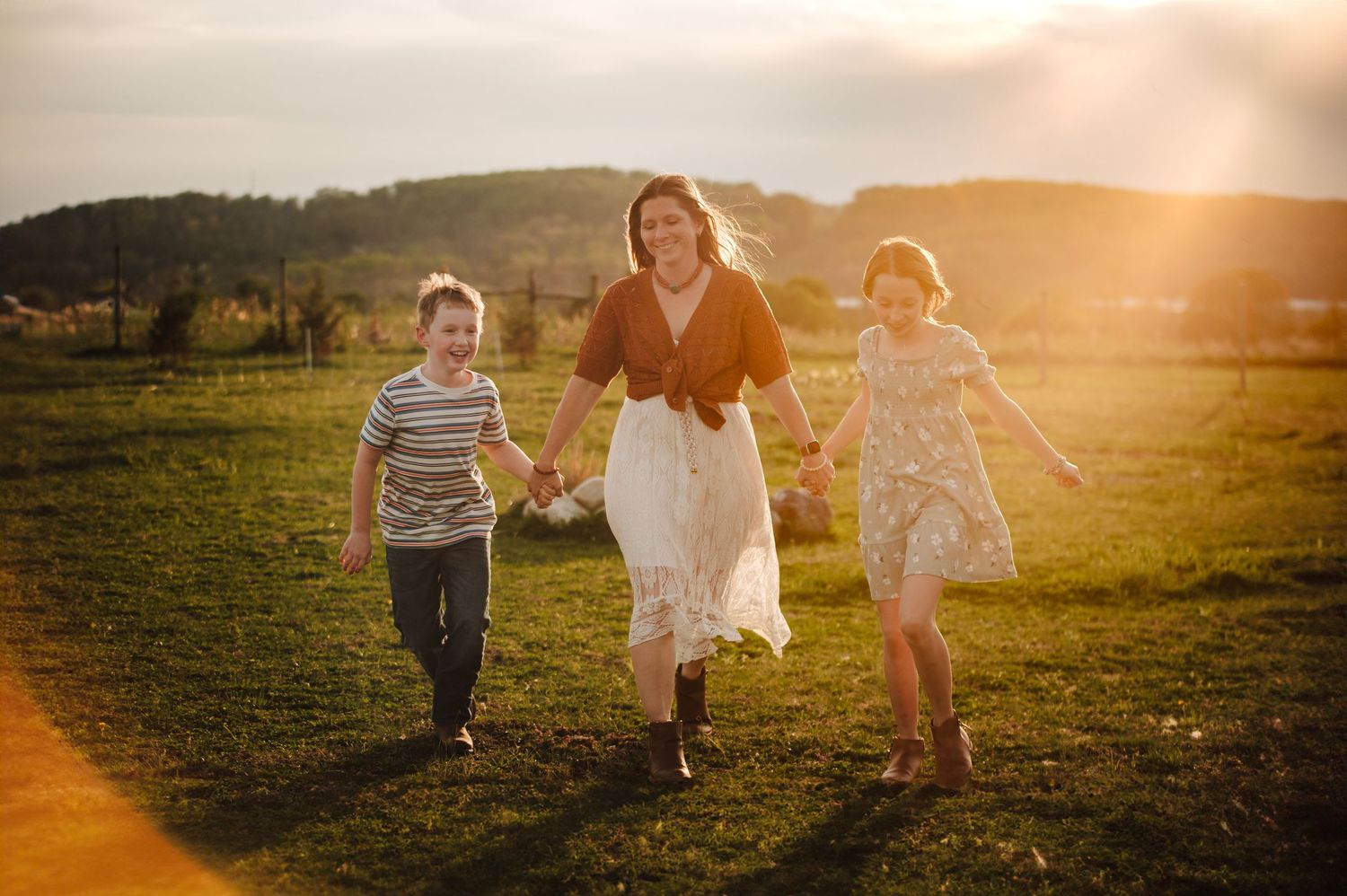 A mother and two children run hand-in-hand through a sunlit green field at golden hour.