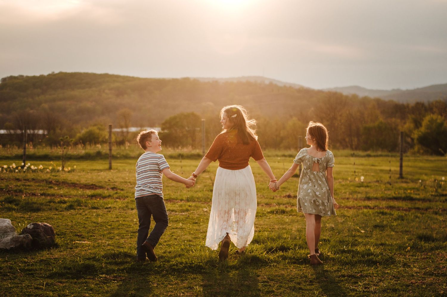 A mother holds hands with her two children walking through a sunlit green field with rolling hills at golden hour.