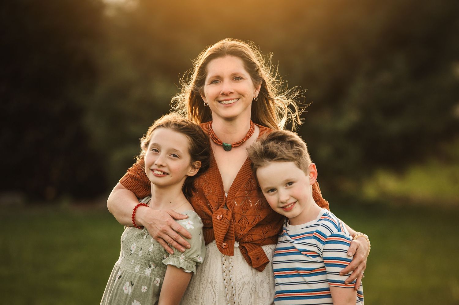 A smiling mother embraces her two young children outdoors in a sunny green field during golden hour.