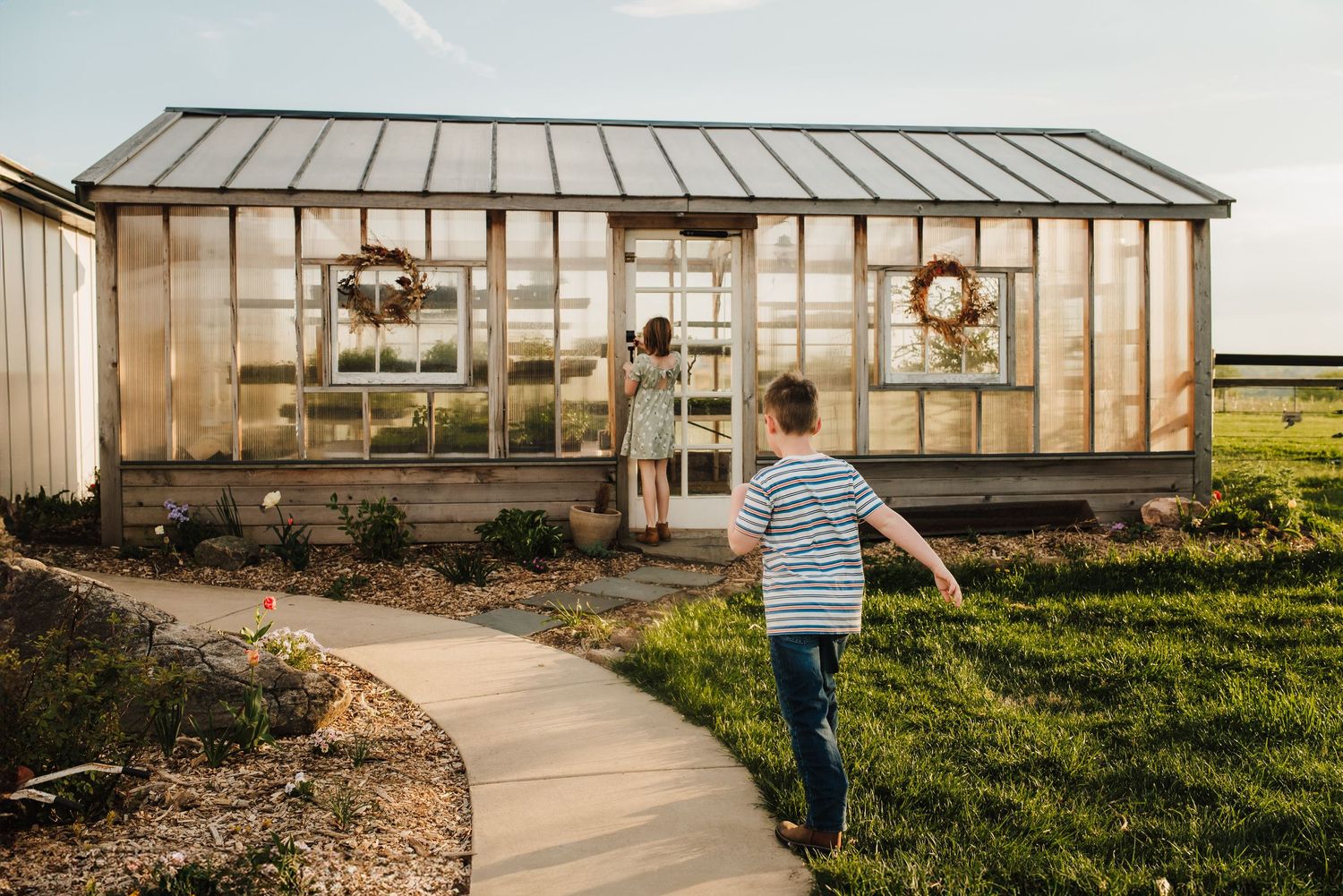 Boy running toward a glass greenhouse with wreaths on the doors, surrounded by a curved path and green lawn at golden hour.
