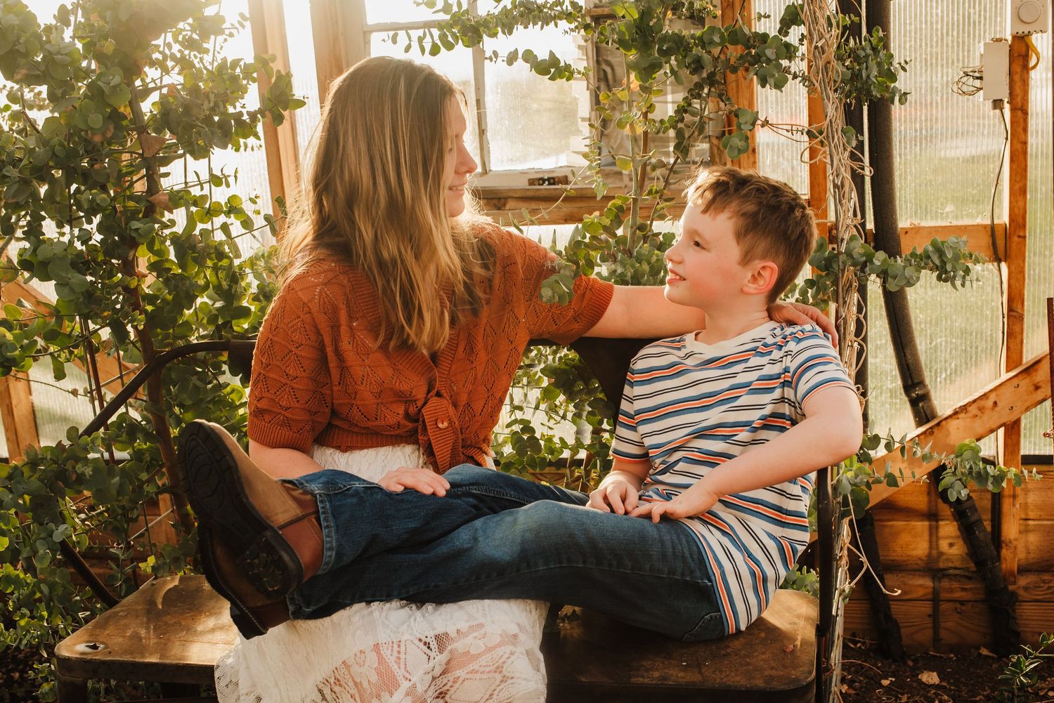 A mother and young boy share a warm moment on a wooden swing surrounded by lush greenery indoors.