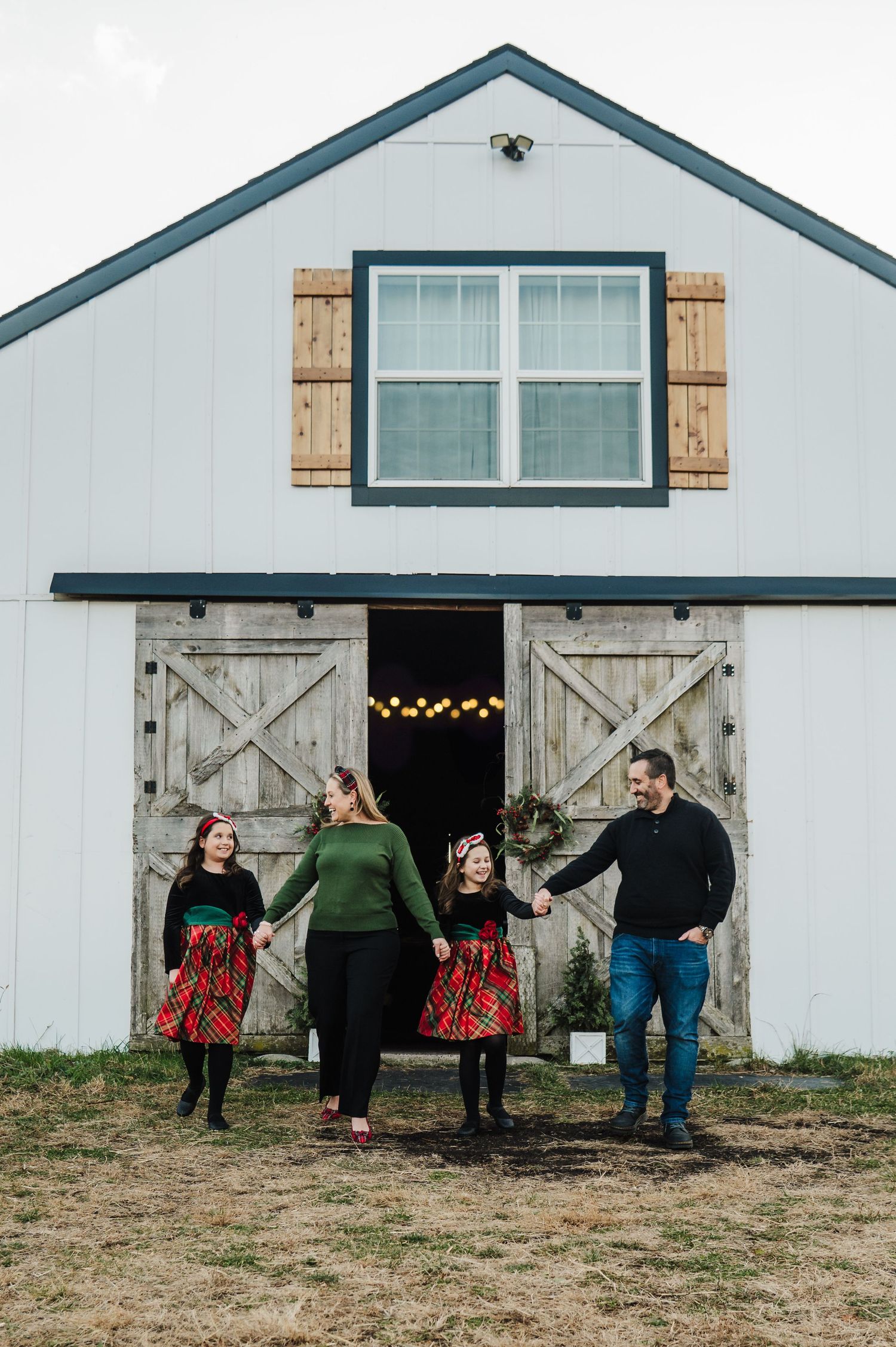 Family of four holding hands in front of a white barn with wooden doors, wearing festive holiday outfits.