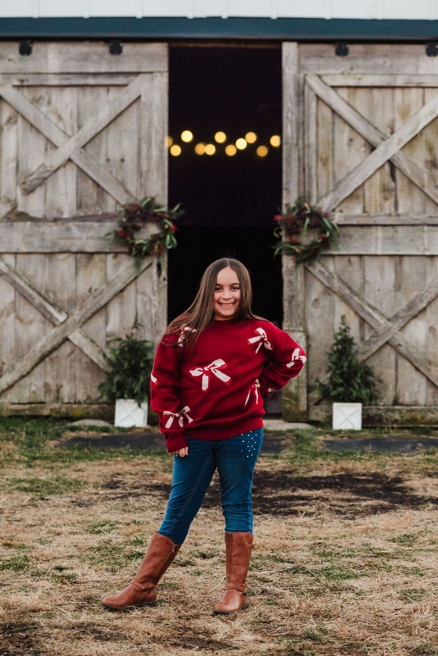 Girl in red bow sweater, jeans, and brown boots poses before a rustic barn decorated with Christmas lights and wreaths.