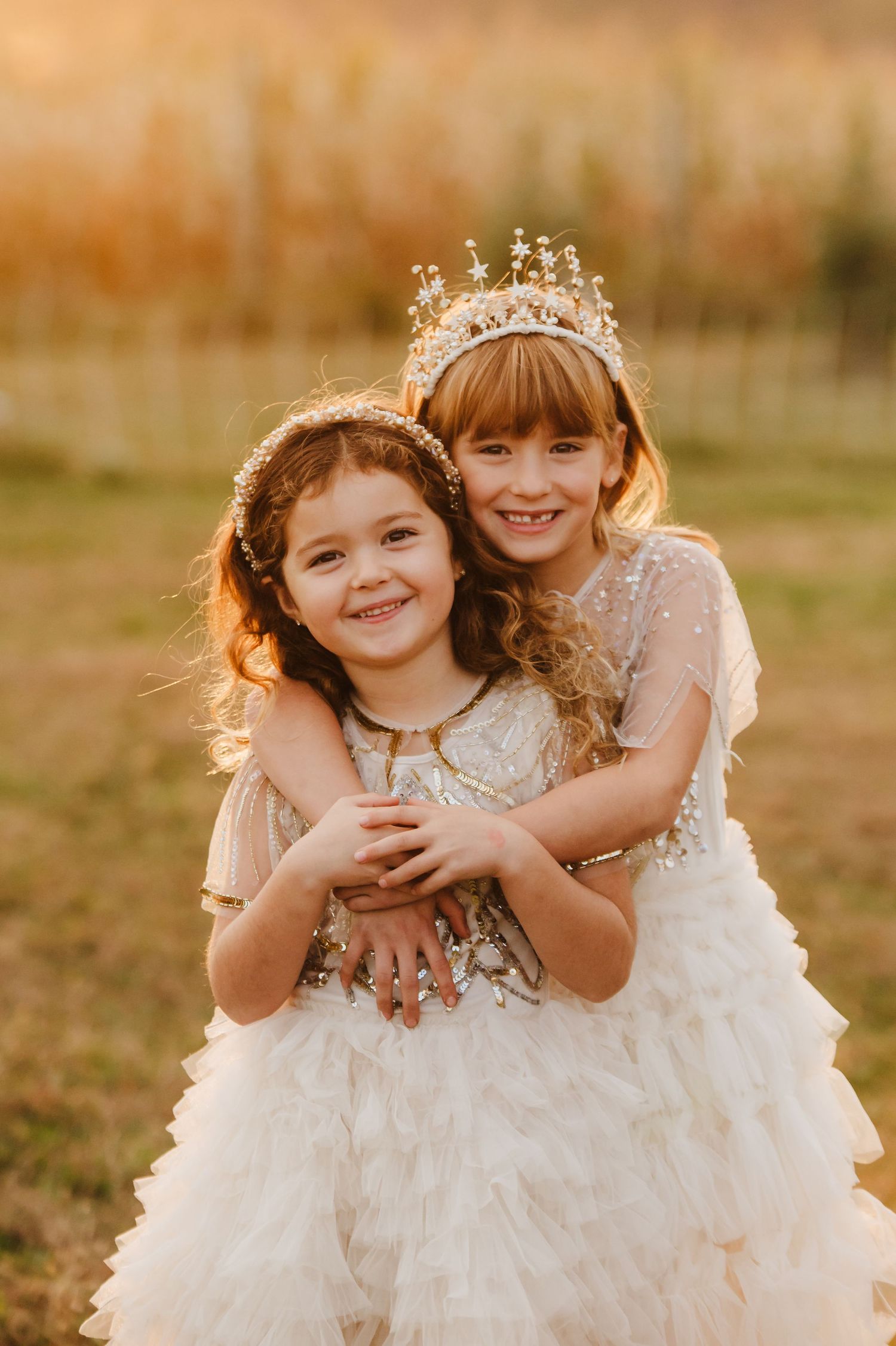Two young girls in white ruffled dresses and gold crowns smile warmly during a golden hour outdoor photo session.