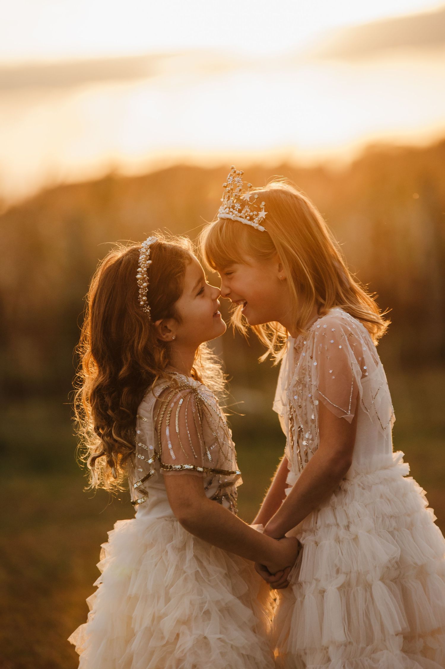 Two little girls in white tulle dresses and tiaras share a sweet moment at golden hour sunset.