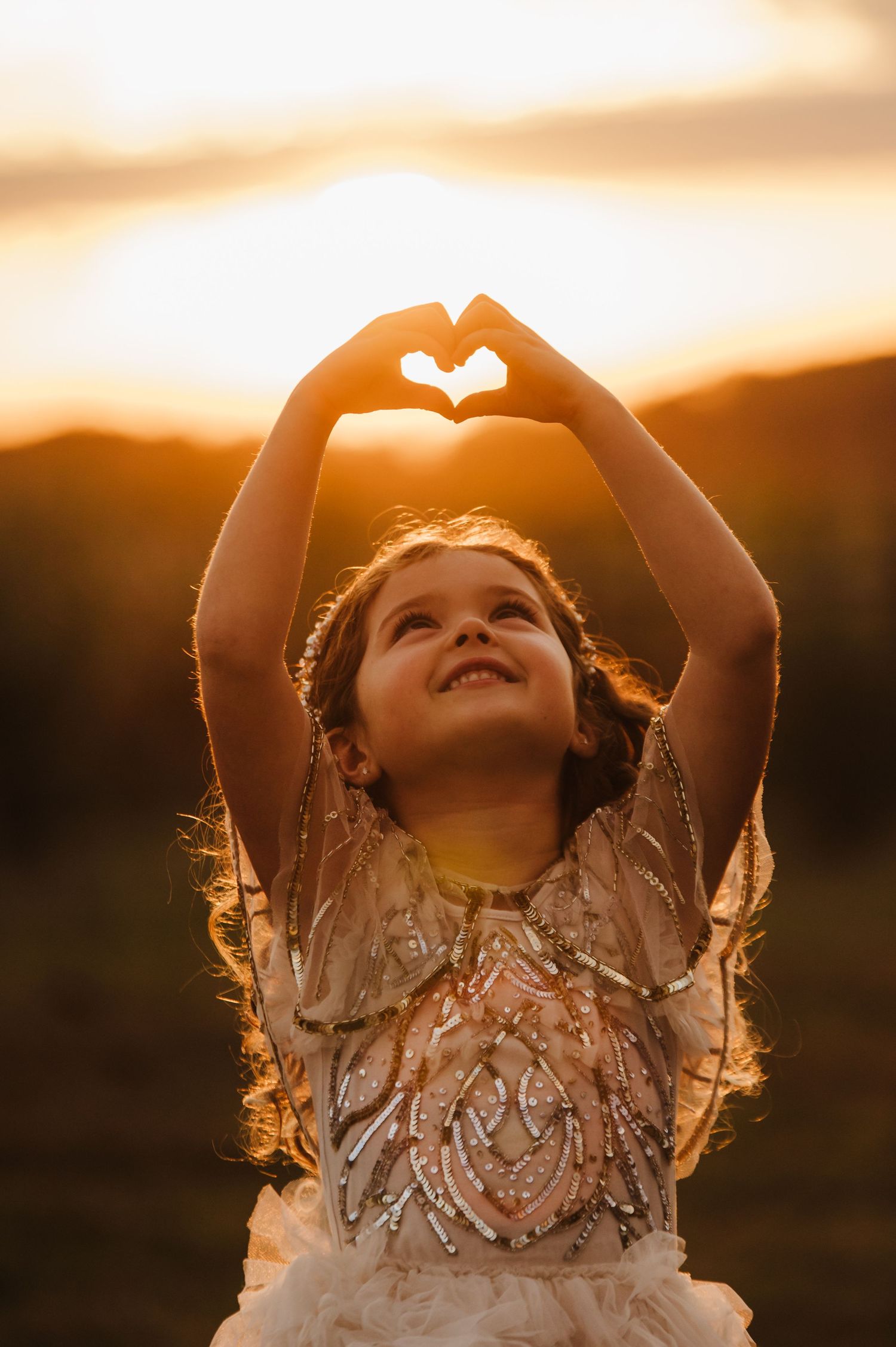 A joyful little girl makes a heart shape with her hands against a golden sunset sky.