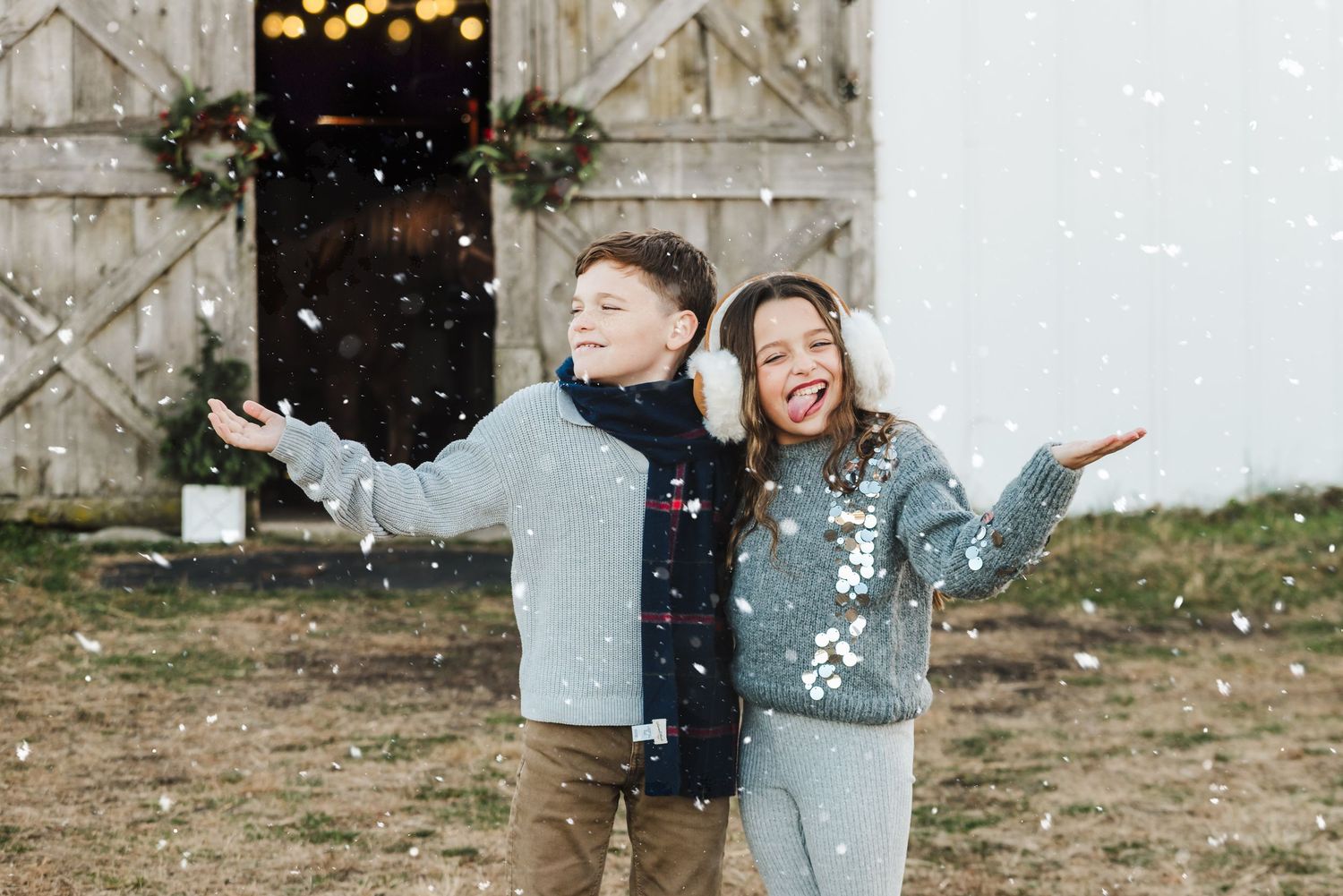 Two children playing in snow near a decorated barn, wearing winter clothes, catching snowflakes outdoors.