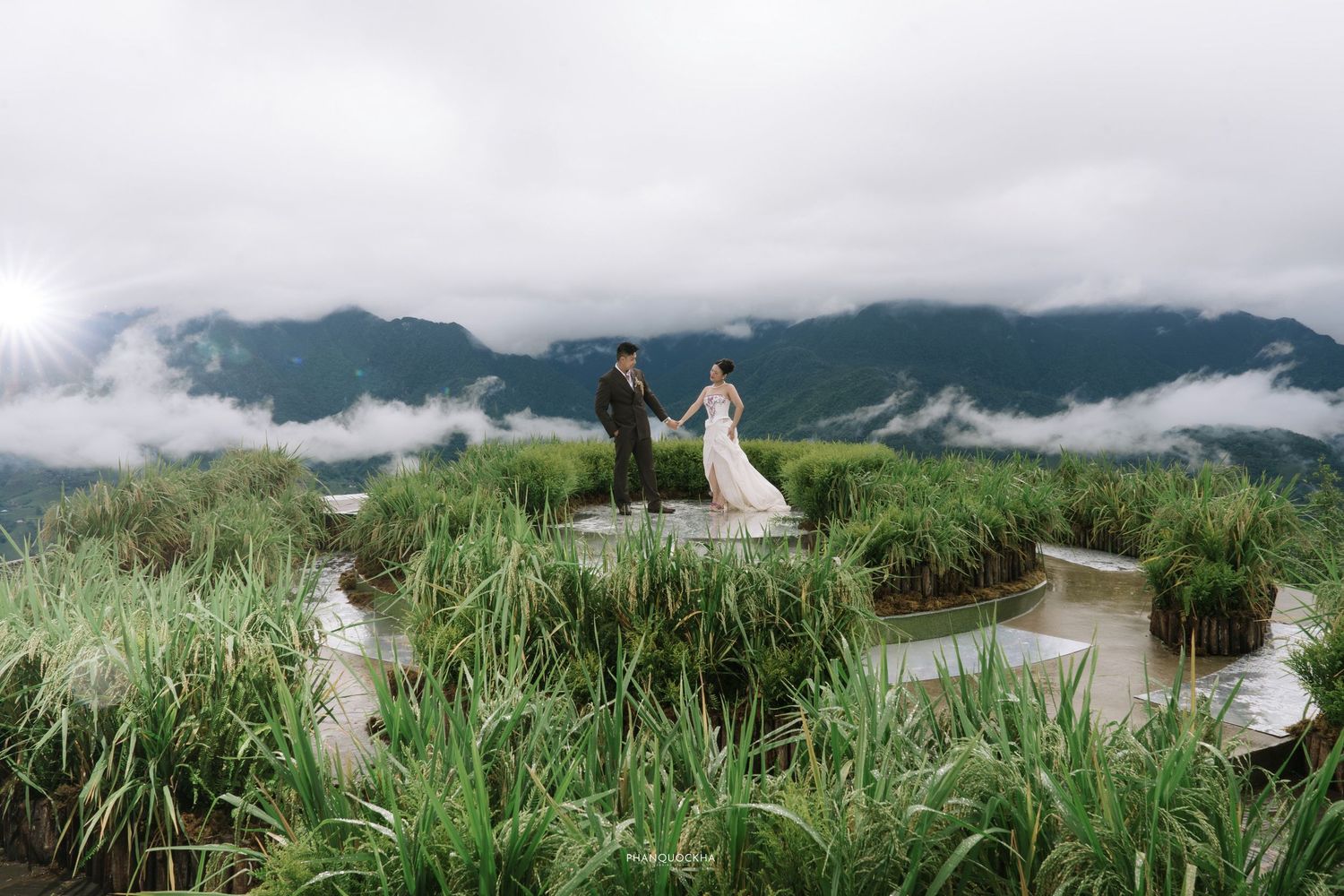 Bride and groom hold hands on a scenic circular terrace surrounded by rice paddies and cloudy mountain landscape.