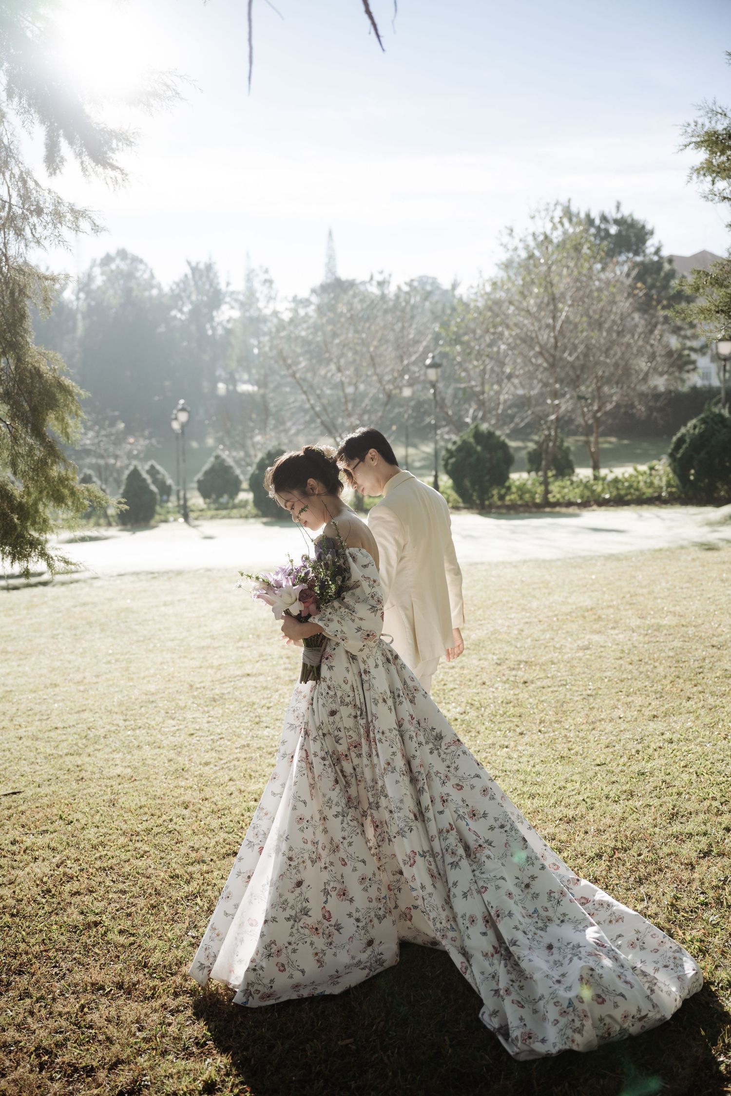 Bride in floral gown and groom in cream suit embracing in sunlit garden with bouquet.