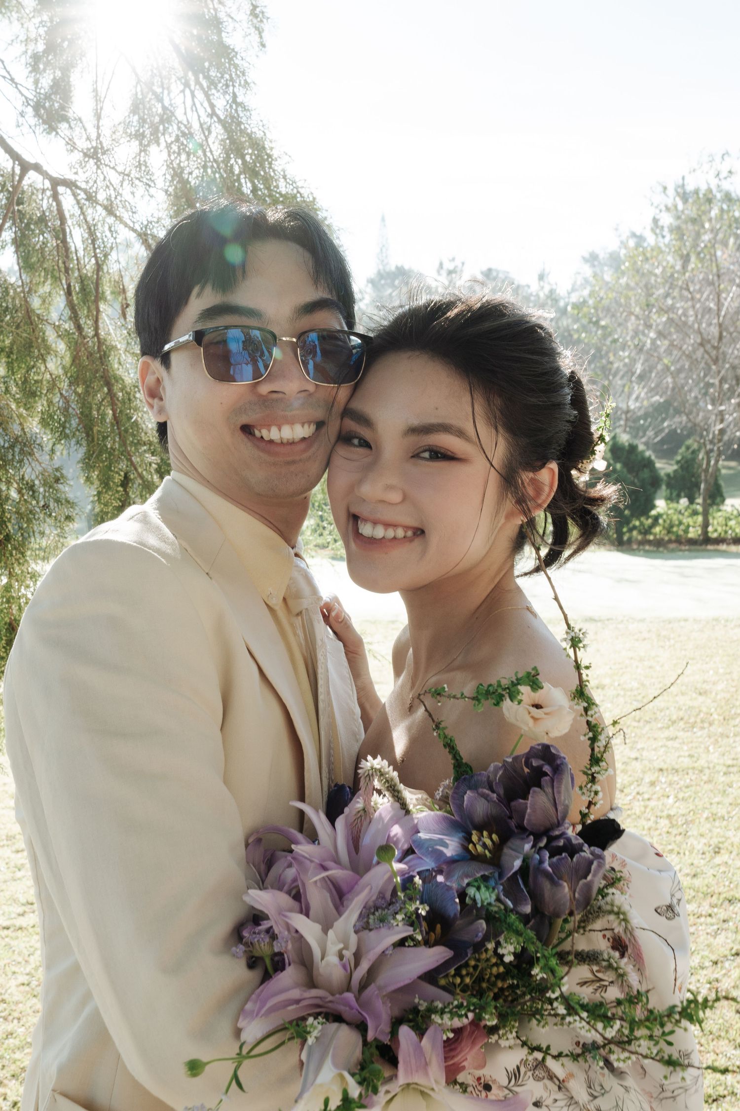 Smiling Asian couple in garden, groom in cream suit with sunglasses, bride holding purple floral bouquet.