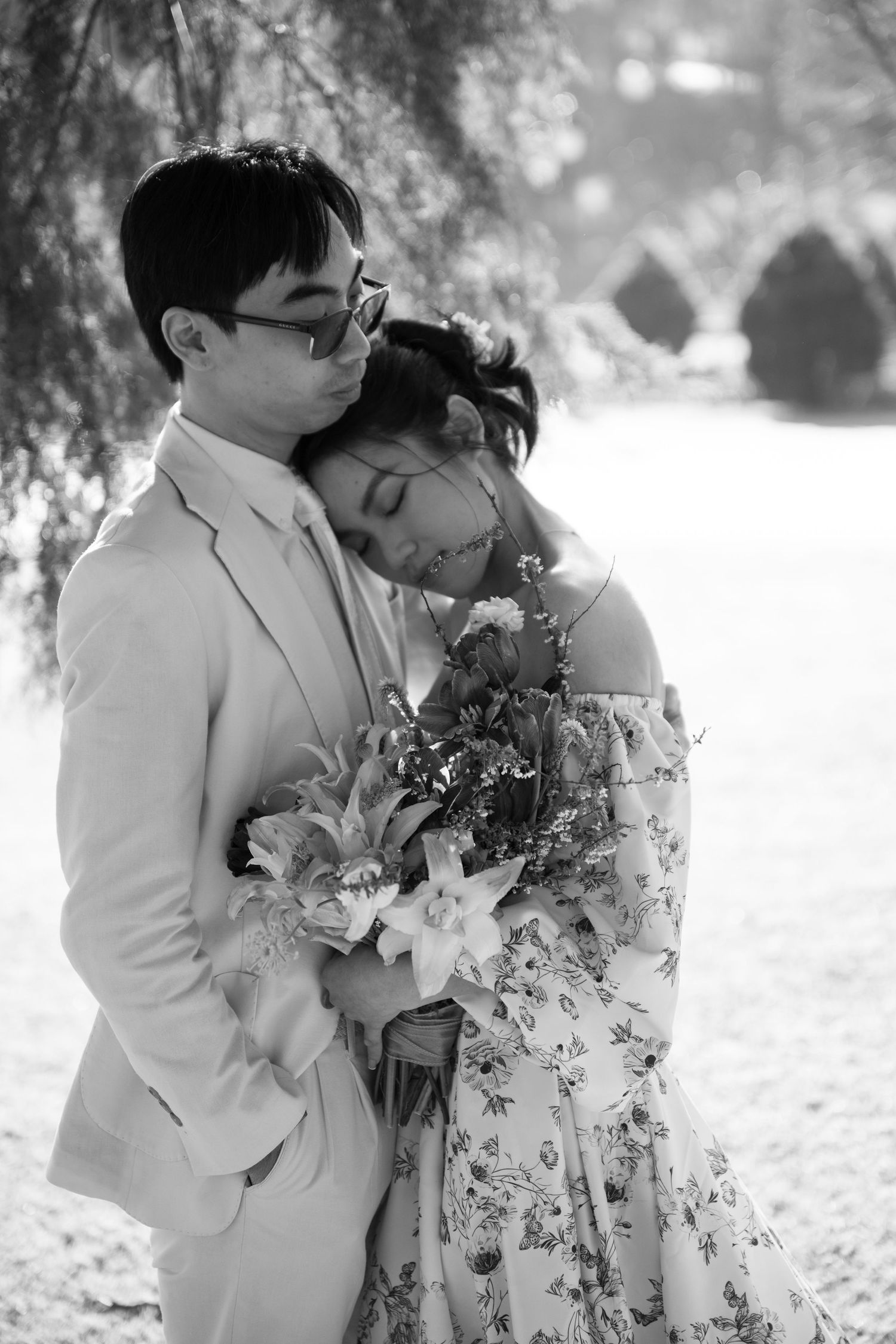 Black and white romantic portrait of couple embracing, bride in floral dress holding bouquet in garden.
